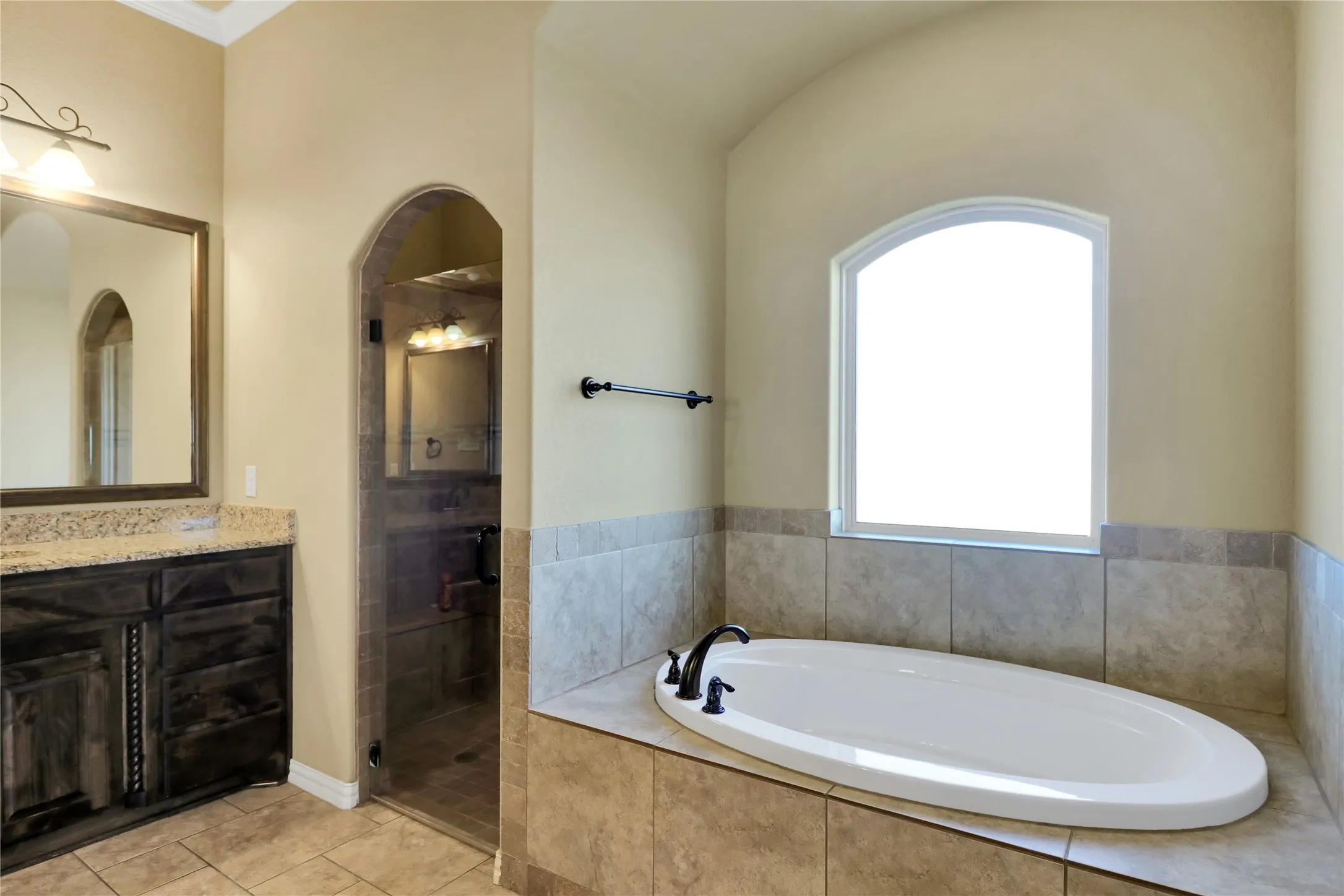Soaking tub with a dark bronze faucet, arched window, tile surround, and a glass-enclosed shower