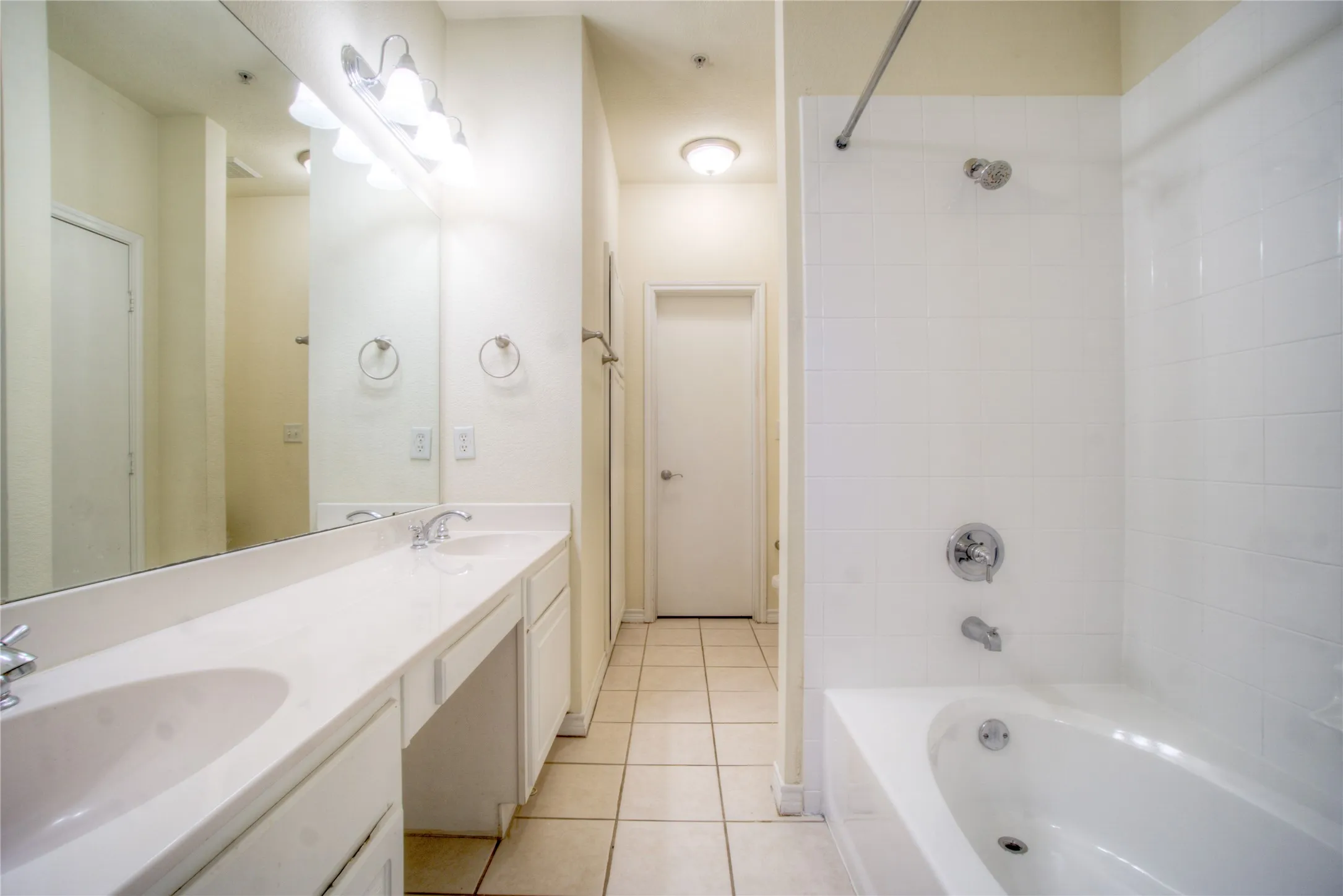 Dual vanity with integrated sinks, full-width mirror, white tile flooring, and white tiled shower/tub combination