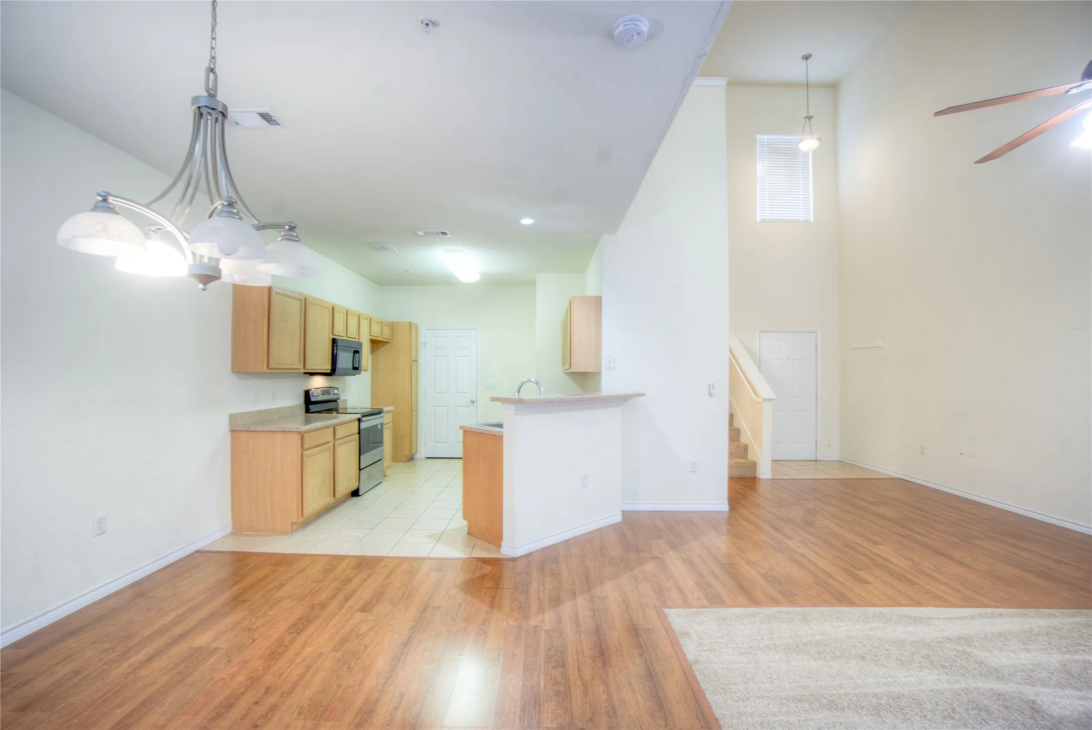 Open-concept living area with wood-finish flooring, a kitchen featuring light wood cabinetry, and a double-height ceiling in the living space