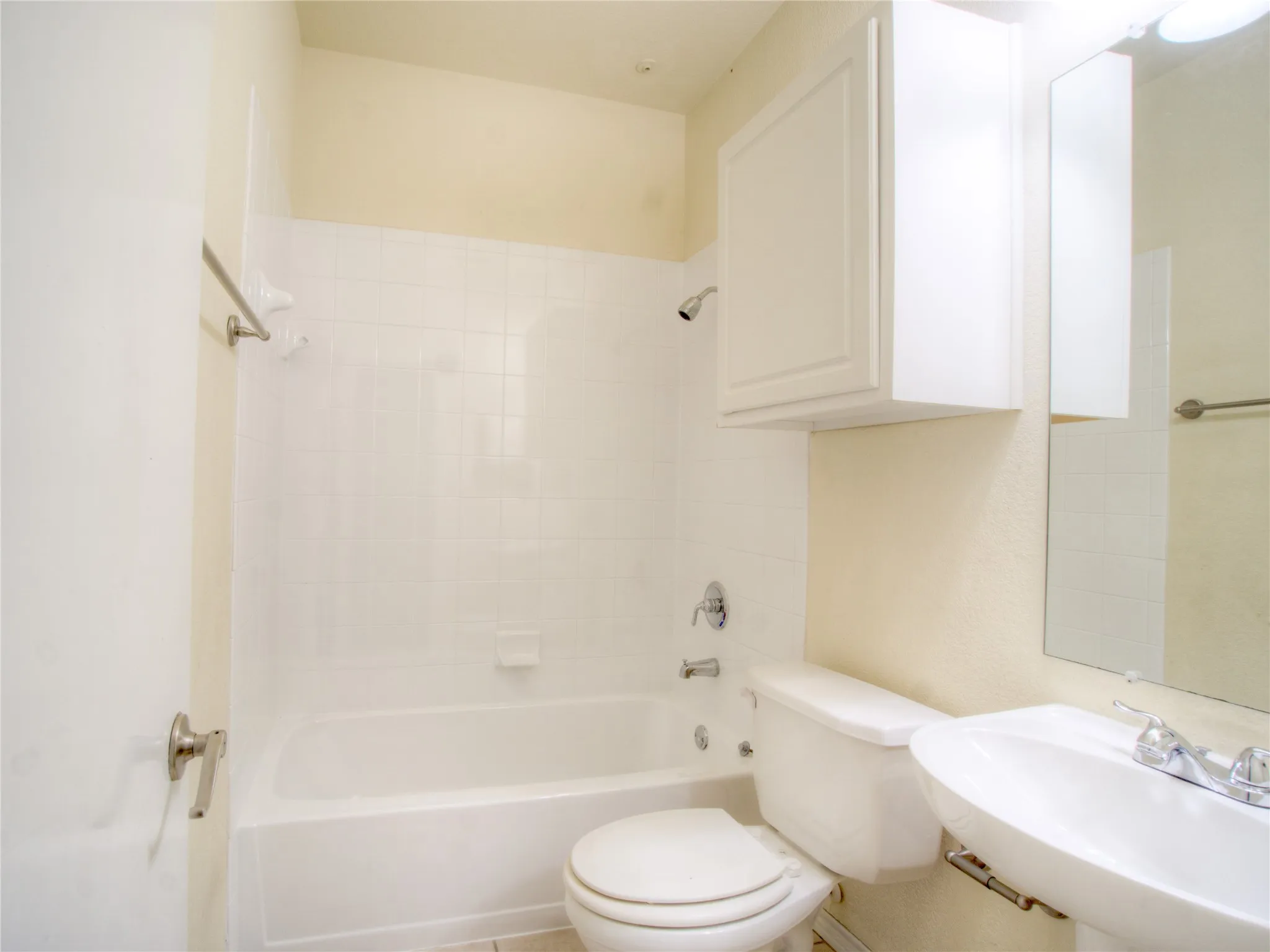 Bathtub with white tile surround, pedestal sink, wall-mounted mirror, and white cabinetry