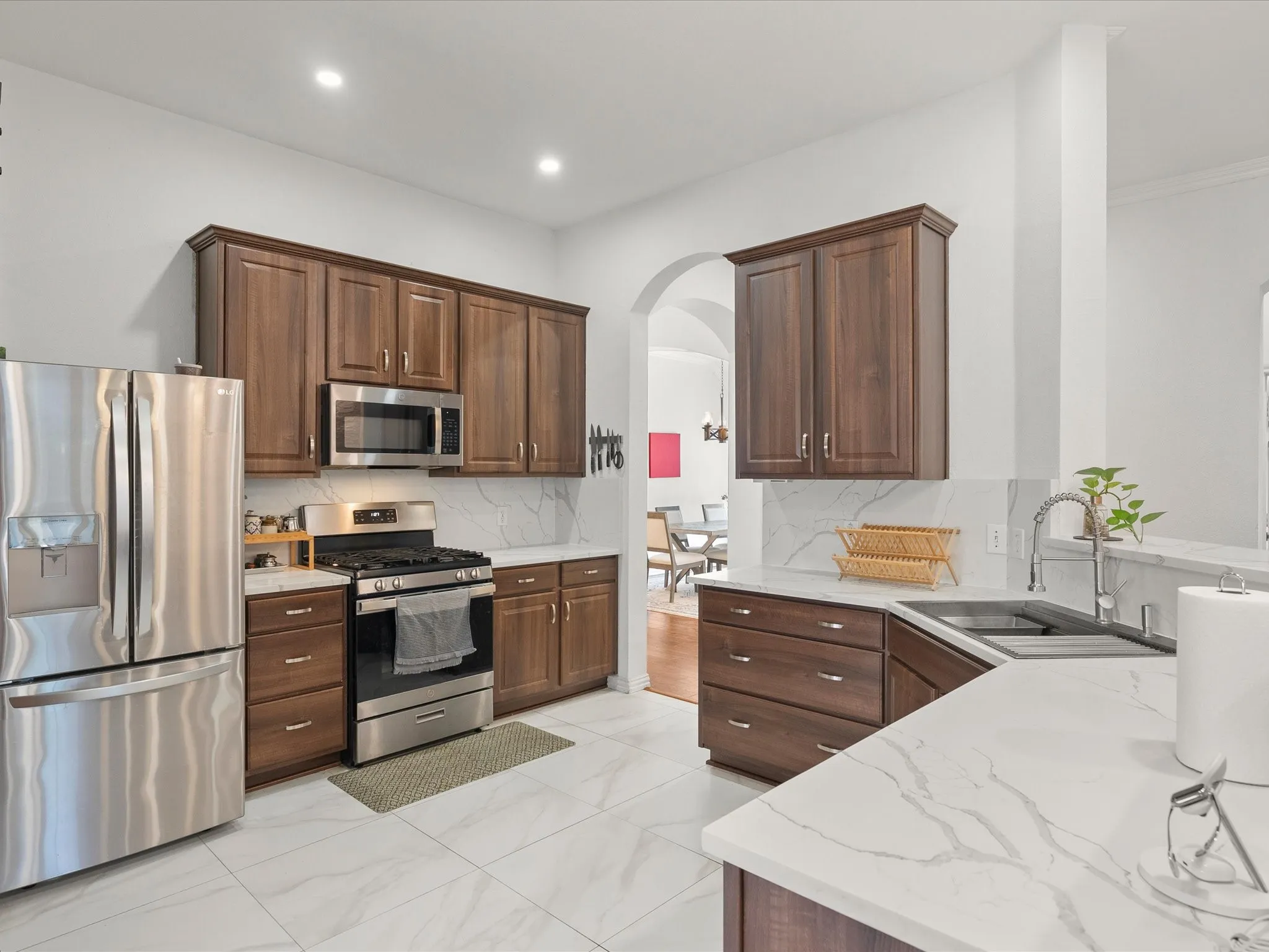 Stainless steel appliances, white patterned countertops, white tile flooring, dark wood-finish cabinetry, and an undermount sink with a gooseneck faucet