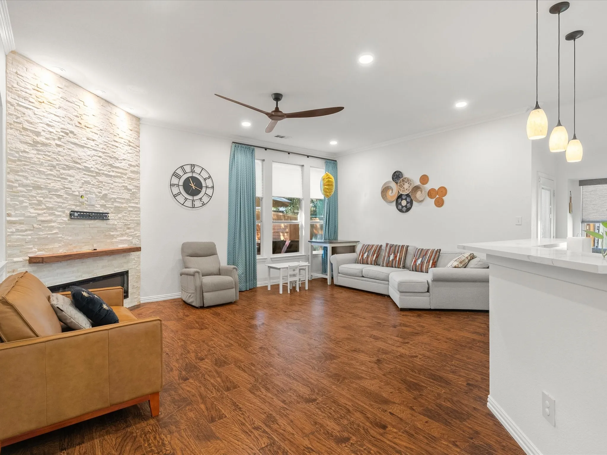 Wood-finish flooring, a stacked stone fireplace with a wood mantle, a ceiling fan, recessed lighting, and pendant lighting