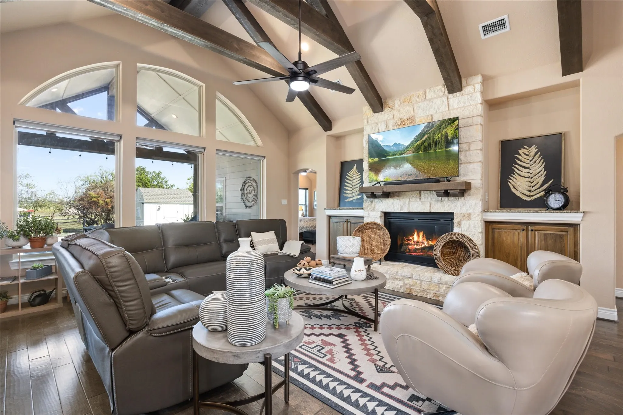 Vaulted ceiling with exposed wood beams, stone-clad fireplace with wood mantle, built-in cabinetry, wood-finish flooring, and large windows