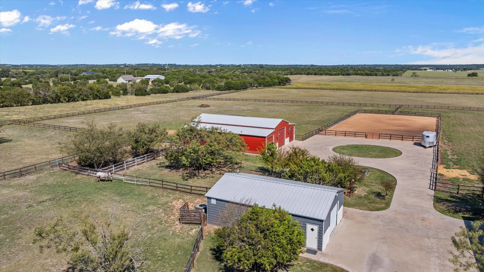 Expansive acreage featuring multiple fenced paddocks, a red barn structure, a separate gray outbuilding, a circular drive, and an outdoor riding arena