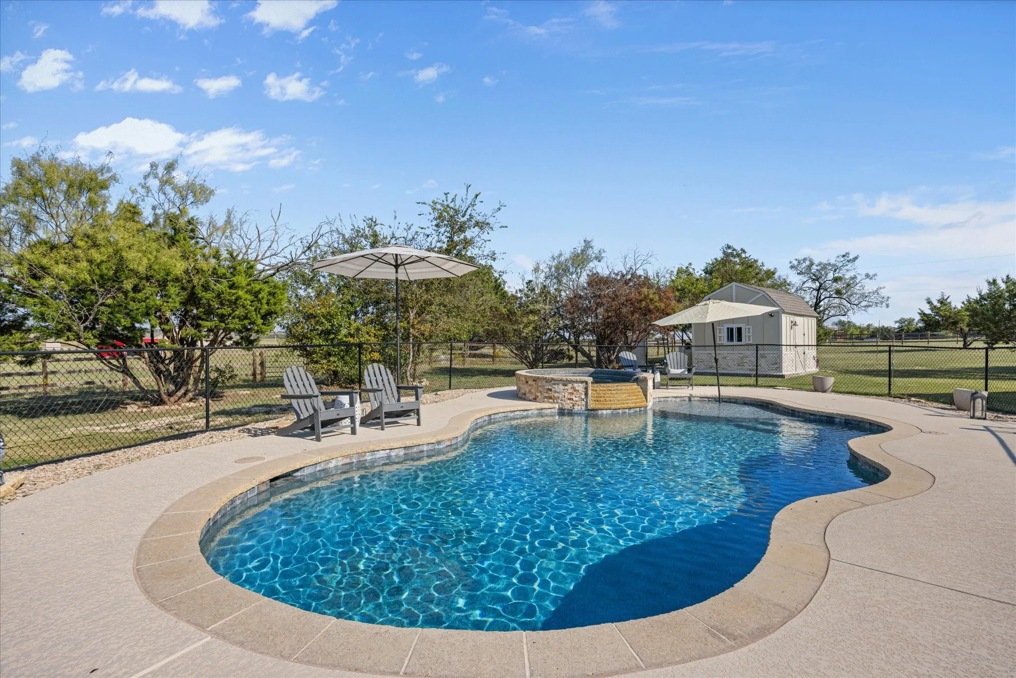 Resort-style backyard oasis featuring a custom free-form saltwater pool with sun shelf, expansive concrete decking, mature landscaping with shade trees, and adjacent poolside cabana