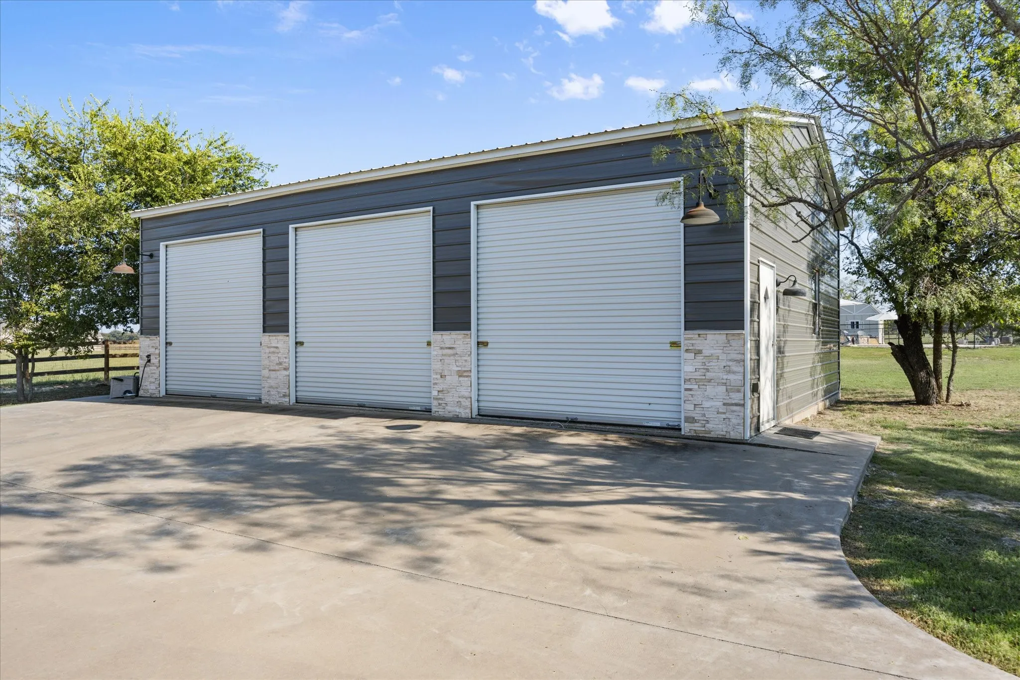 Steel-sided outbuilding featuring three roll-up doors, decorative stone veneer accents, and a concrete driveway