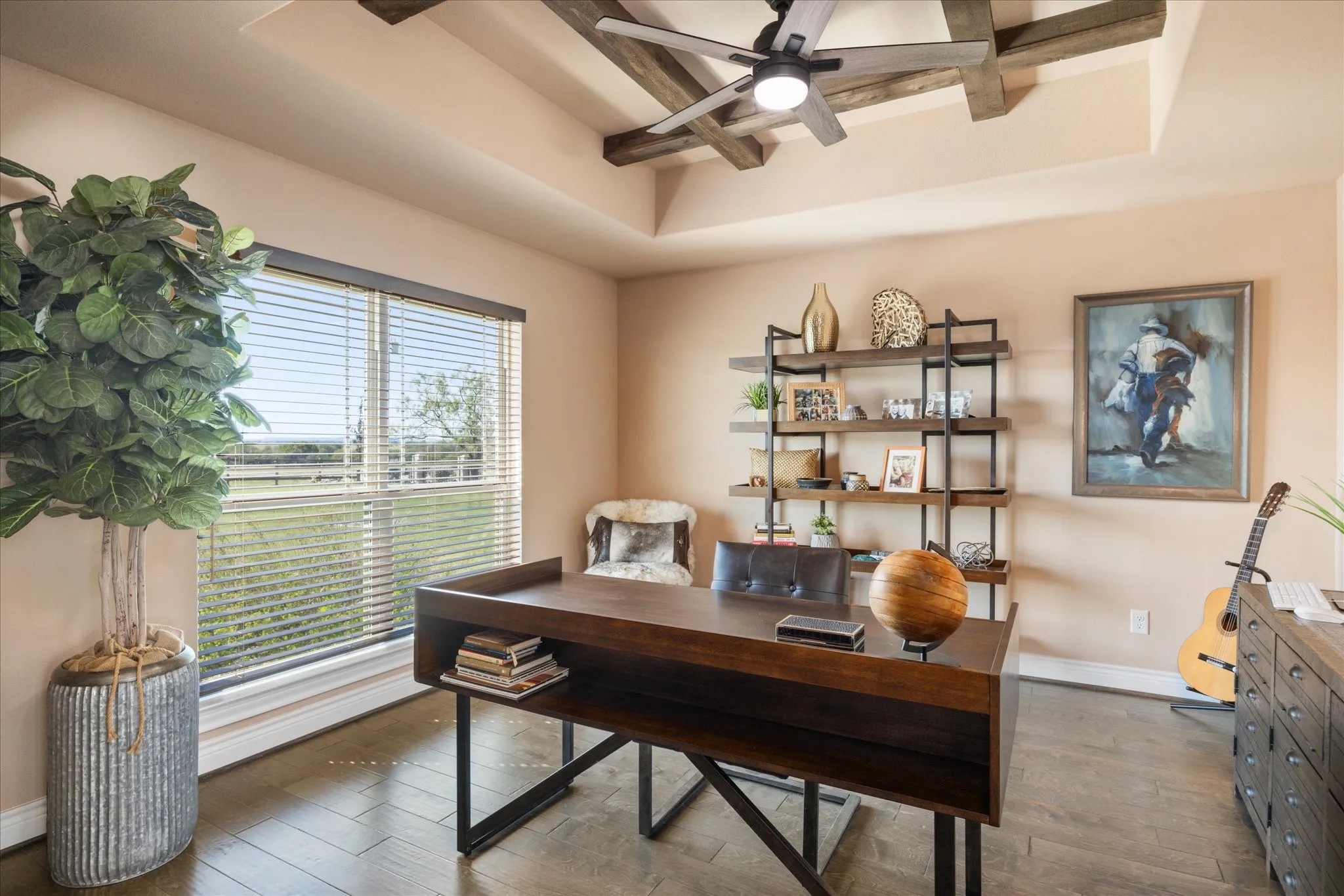 Tray ceiling with exposed wood beams and a ceiling fan, wood-finish flooring, large window with blinds, built-in shelving with dark metal framing, and a wood desk with metal legs