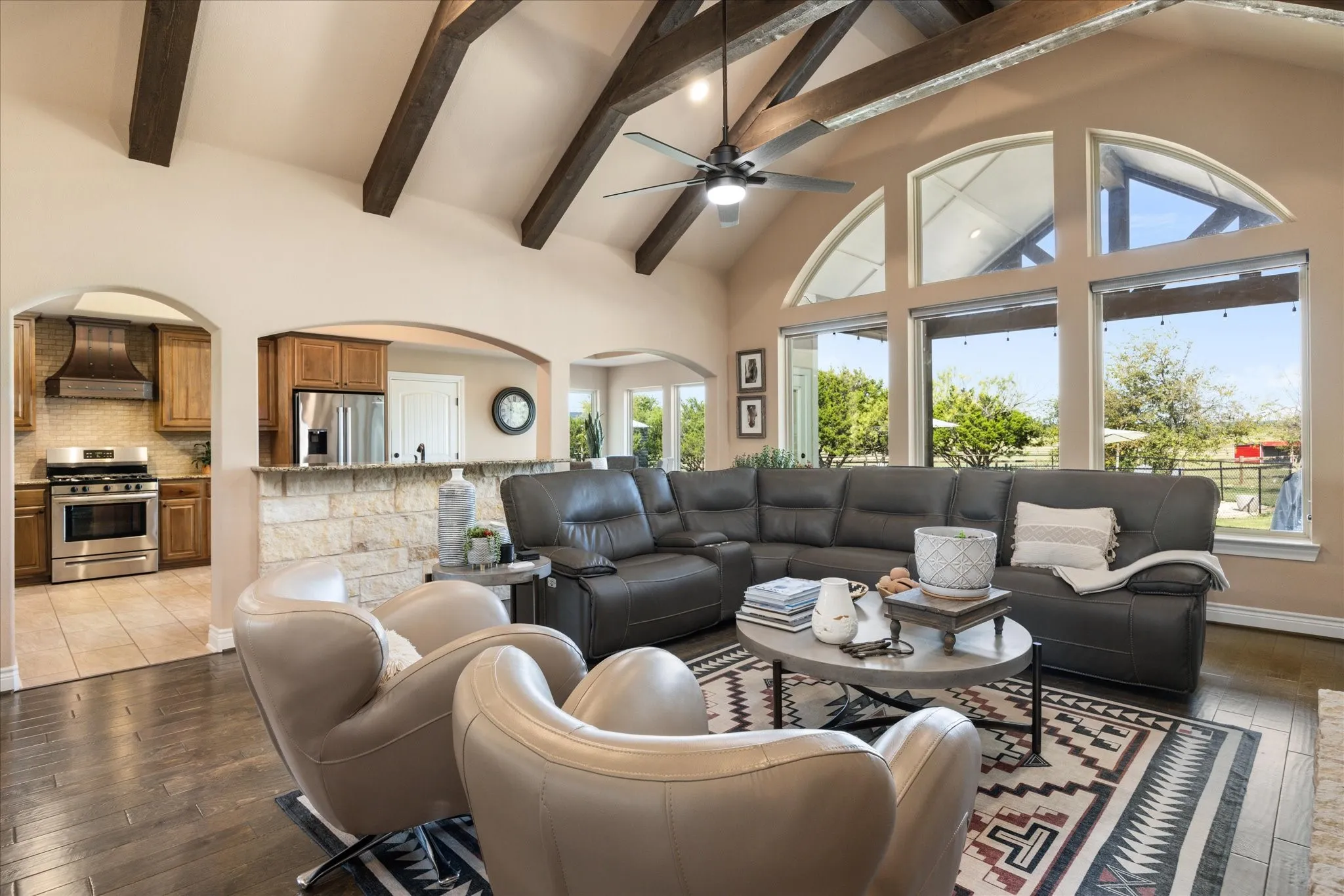 Vaulted ceiling with exposed wood beams, expansive window array with arched transoms, wood-finish flooring, and a stone-clad breakfast bar