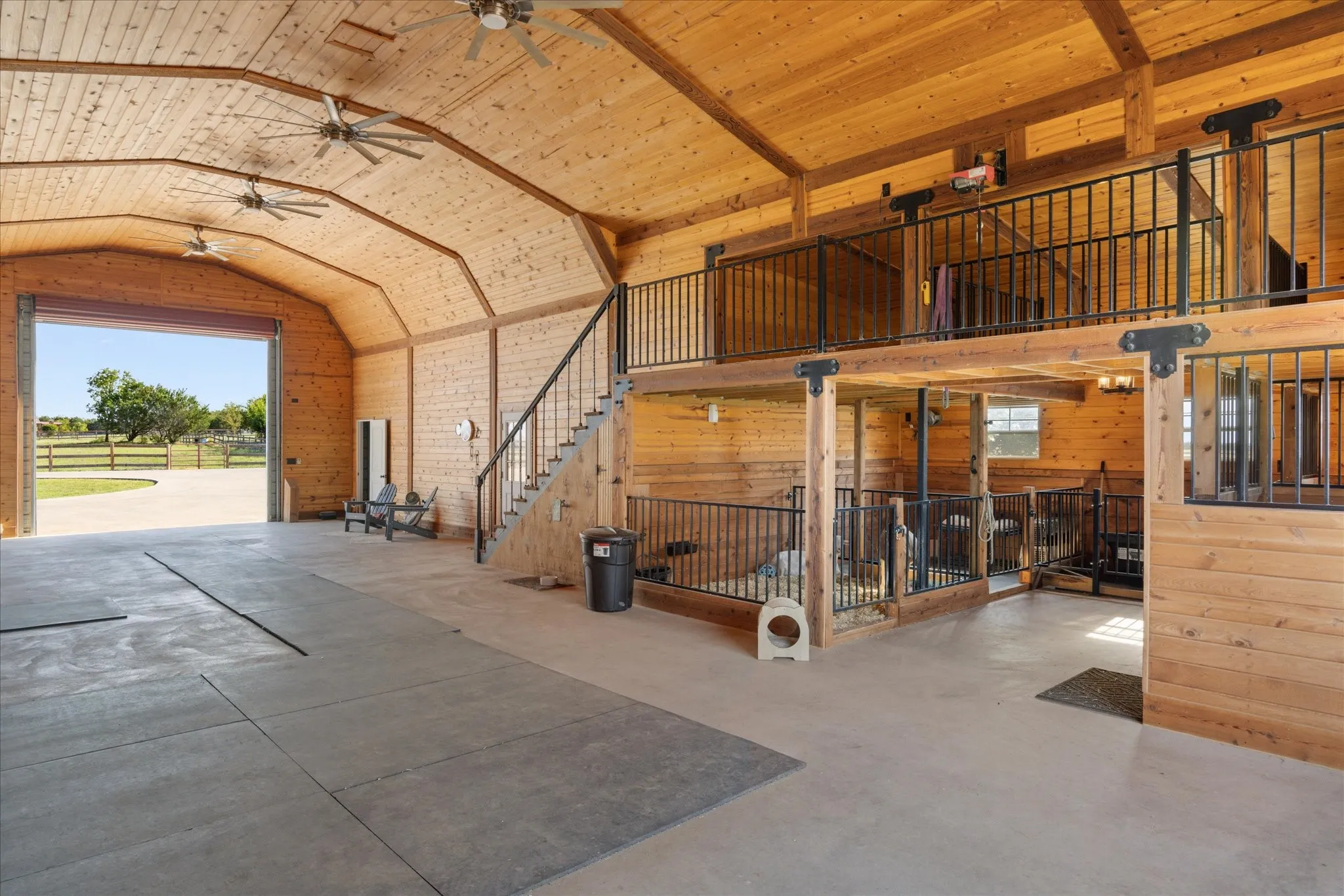 Vaulted wood-paneled ceiling with exposed beams, concrete flooring, dual-level interior with black metal railings, wood staircase, and multiple ceiling fans