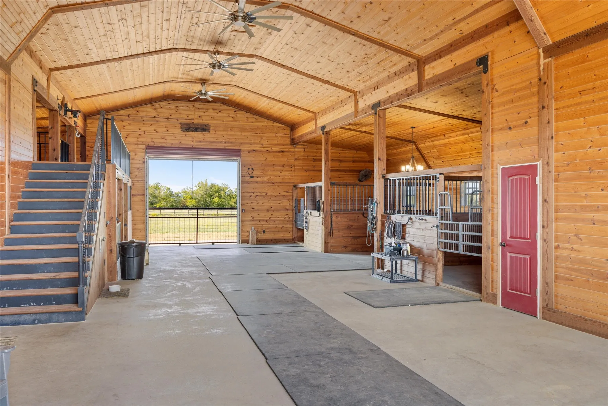 Vaulted wood-paneled ceiling with exposed beams, concrete flooring, interior staircase with metal railing, multiple interior stalls with metal grates, and a roll-up door