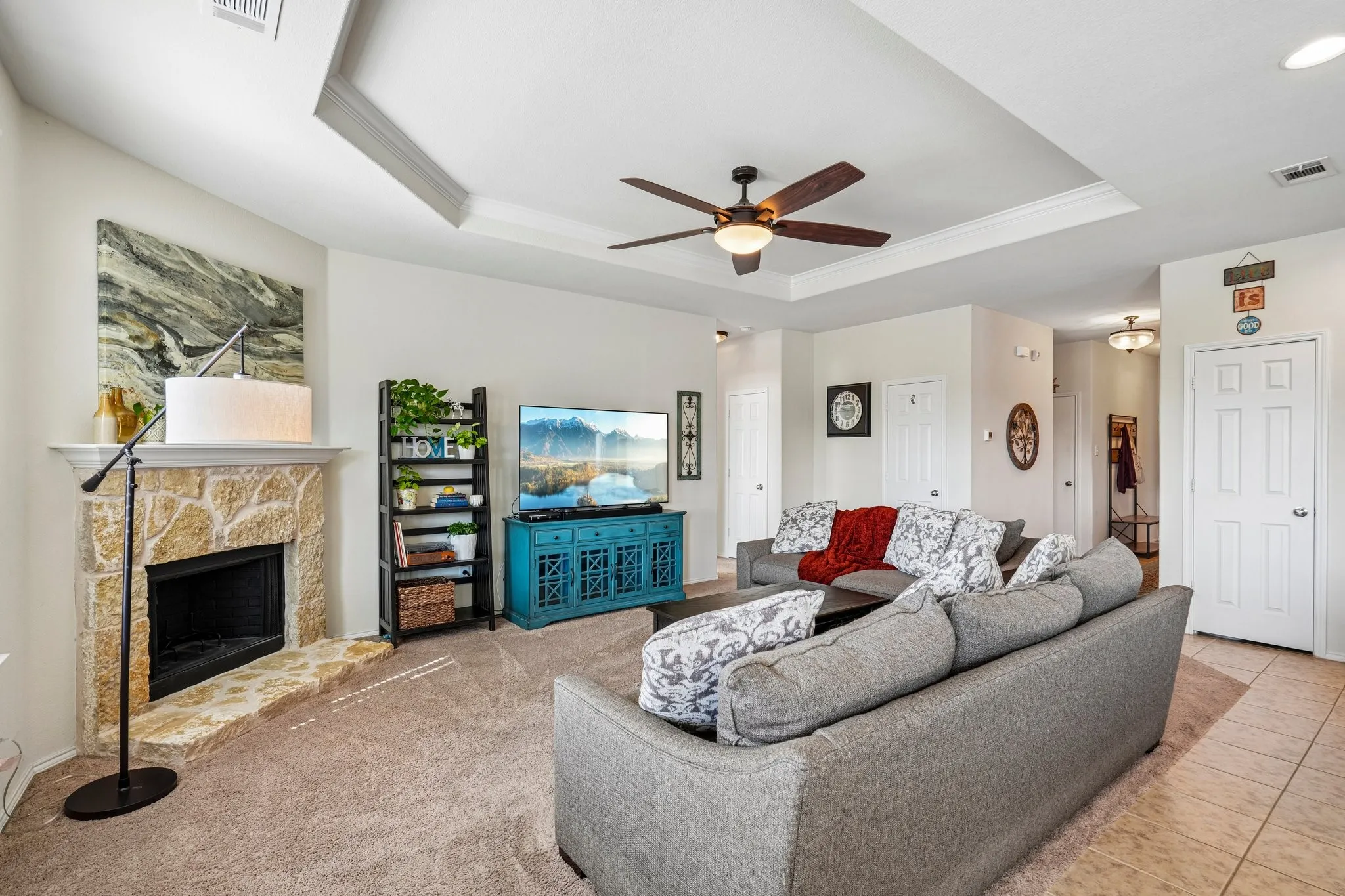 Tray ceiling with a ceiling fan, stone-clad fireplace with mantel, and carpet flooring