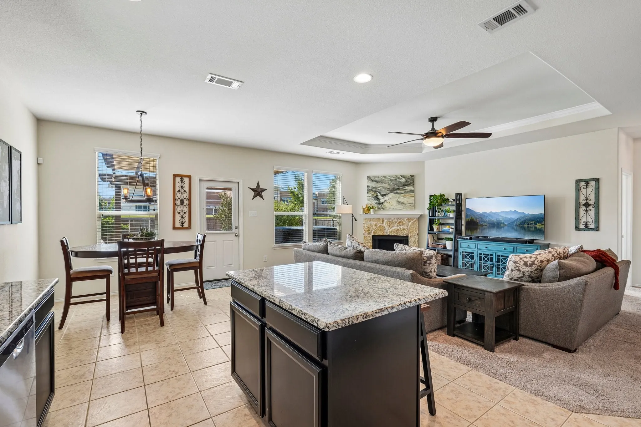 Open-concept floor plan featuring a kitchen island with stone countertop, tile flooring, a fireplace with stone surround, and tray ceilings