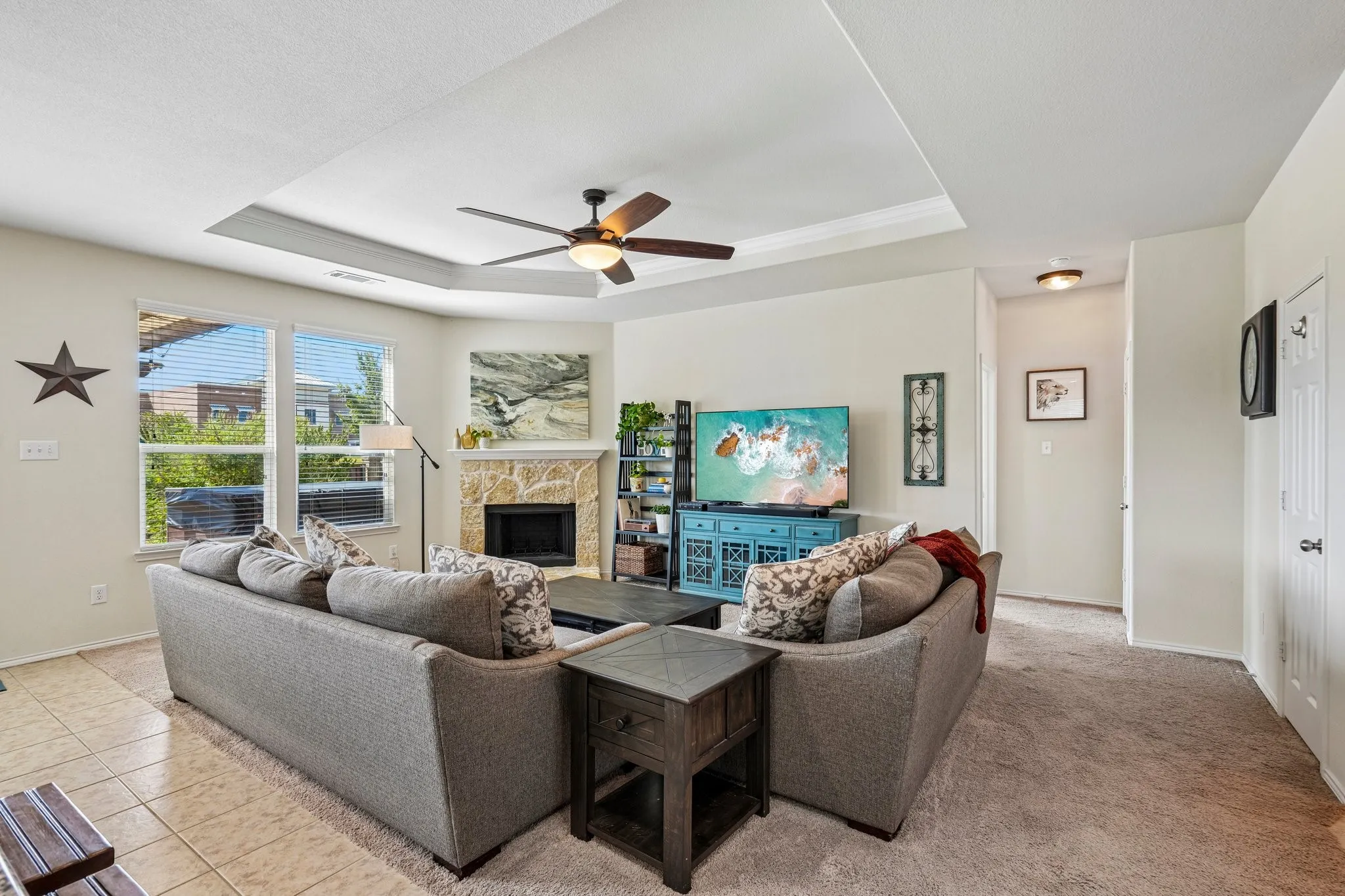 Living room featuring a fireplace with stone surround, tray ceiling, ceiling fan, and multiple windows