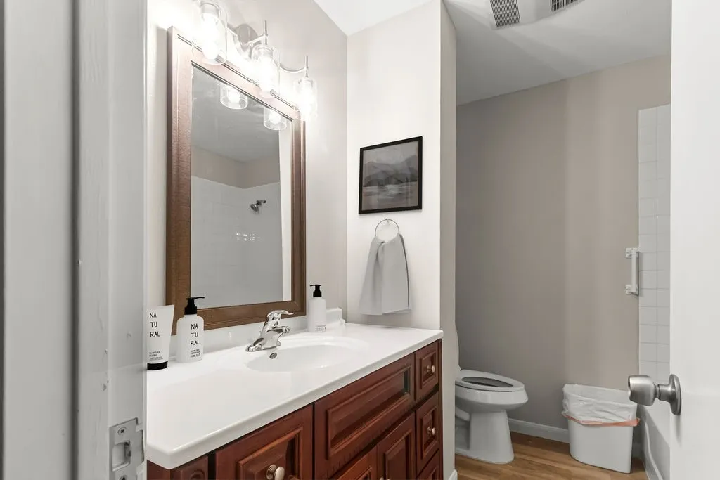 Wood-finish flooring, single vanity with a white countertop, wood-frame mirror, and wall-mounted lighting fixture