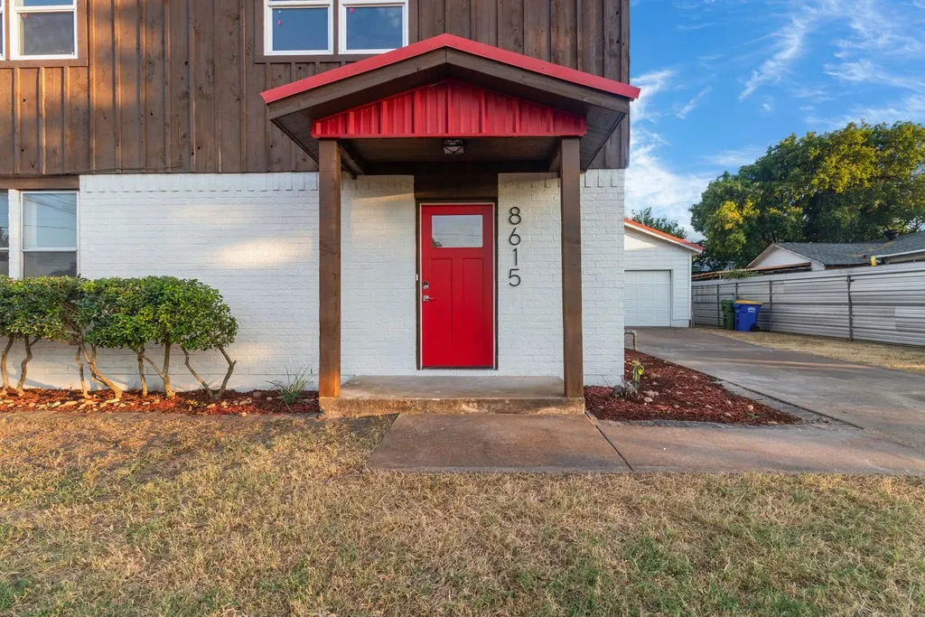 Red front door with vertical house numbers, wood-paneled upper exterior, white brick lower exterior, covered entry, and concrete walkway