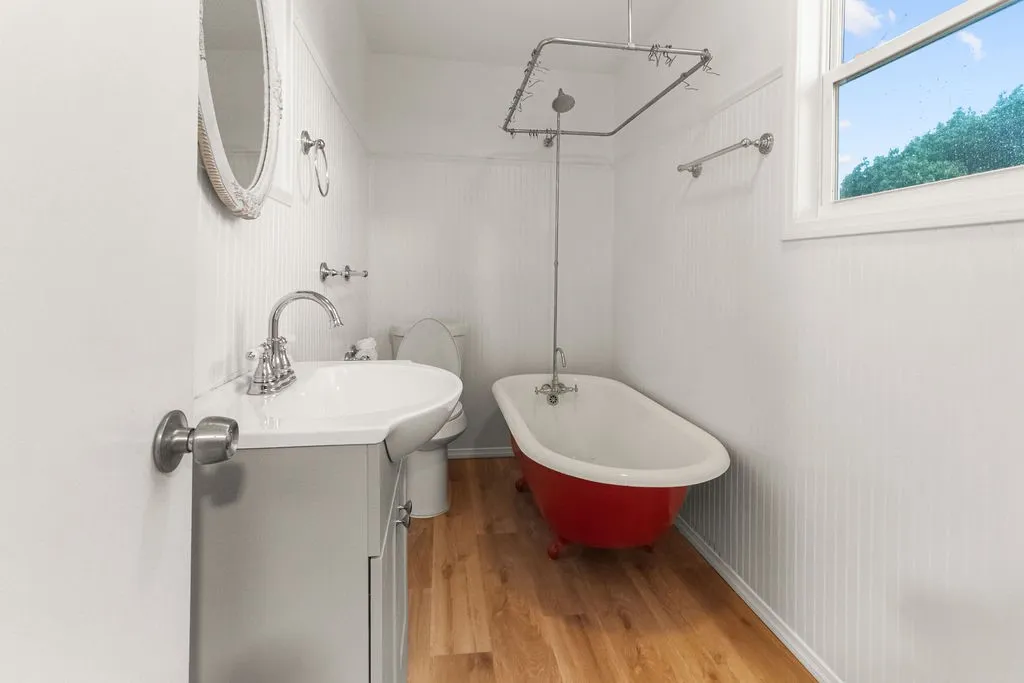 Freestanding red clawfoot tub with shower, white beadboard walls, wood-finish flooring, white vanity with integrated sink, and white-framed window
