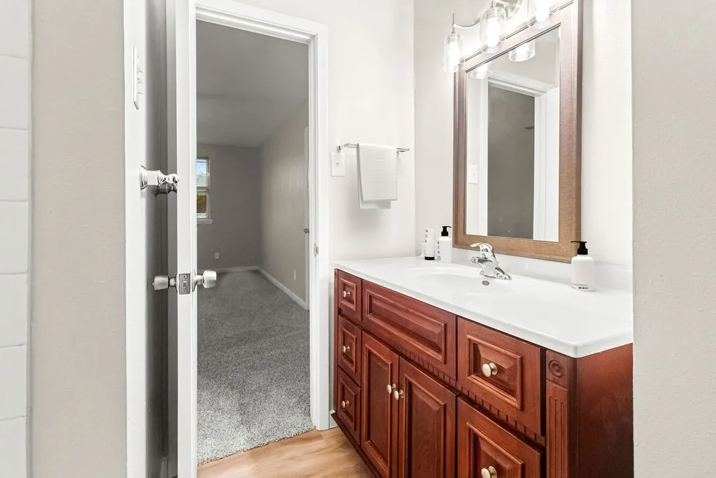 Spa-inspired bathroom featuring warm wood-finish cabinetry with multiple drawers and hardware accents, a white solid-surface countertop with integrated sink, framed mirror with brushed metal trim, and access to an adjoining walk-in closet with gray tile flooring