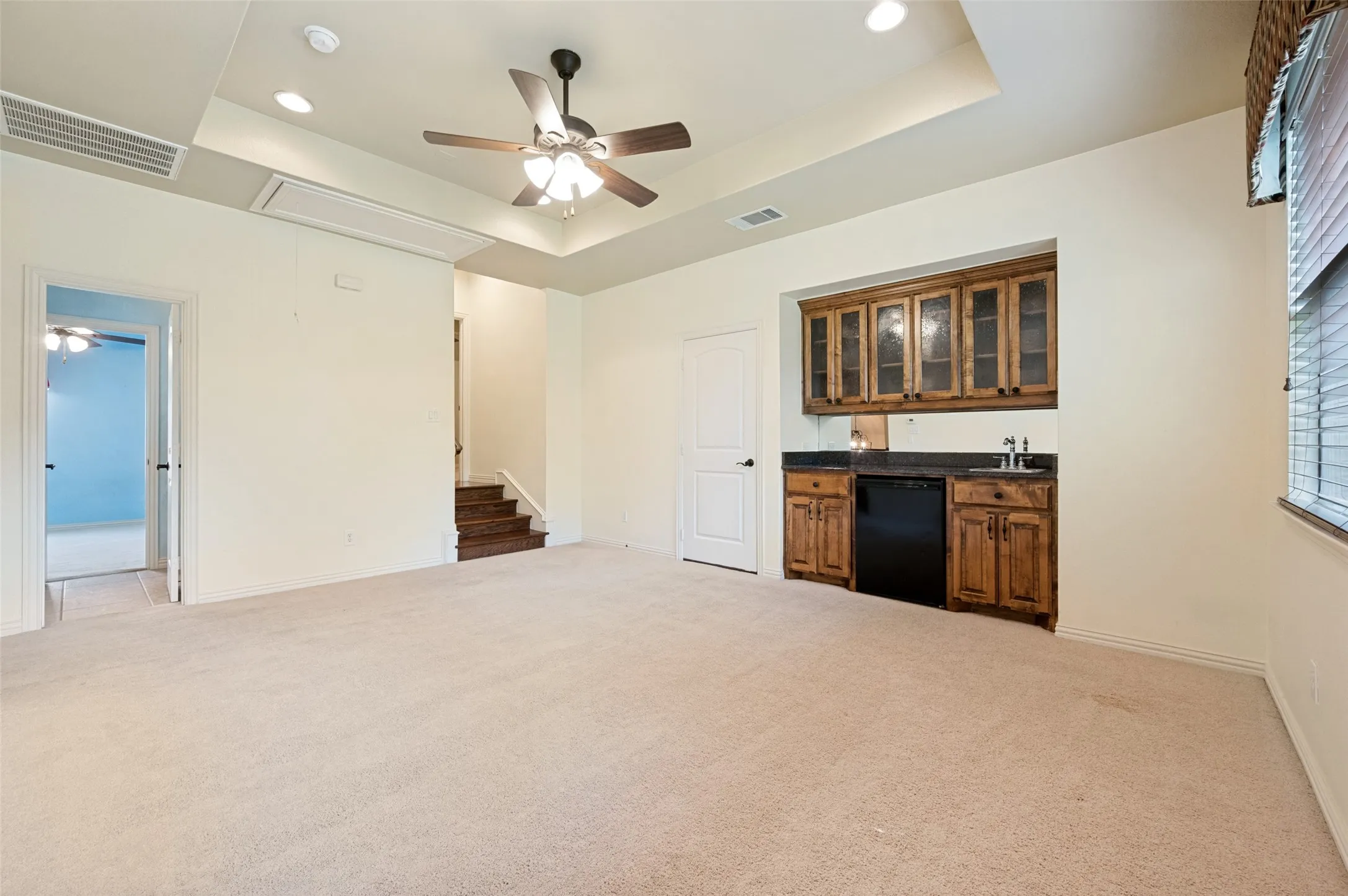 Spacious entertainment area with neutral carpet, tray ceiling, and a built-in wet bar featuring wood cabinetry, a sink, and a compact refrigerator