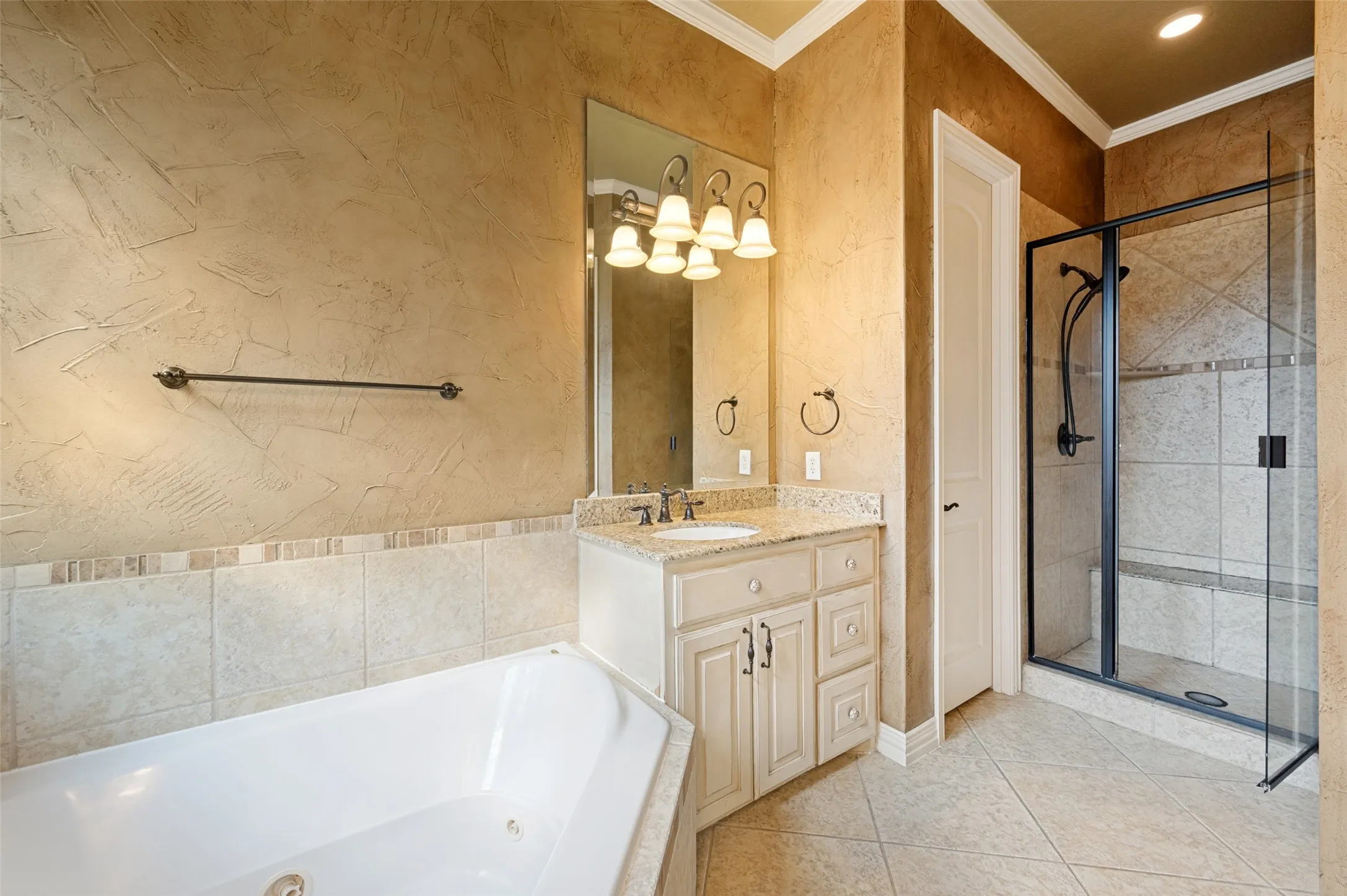 Corner soaking tub, separate glass-enclosed shower with tile surround, single vanity with countertop, and tile flooring