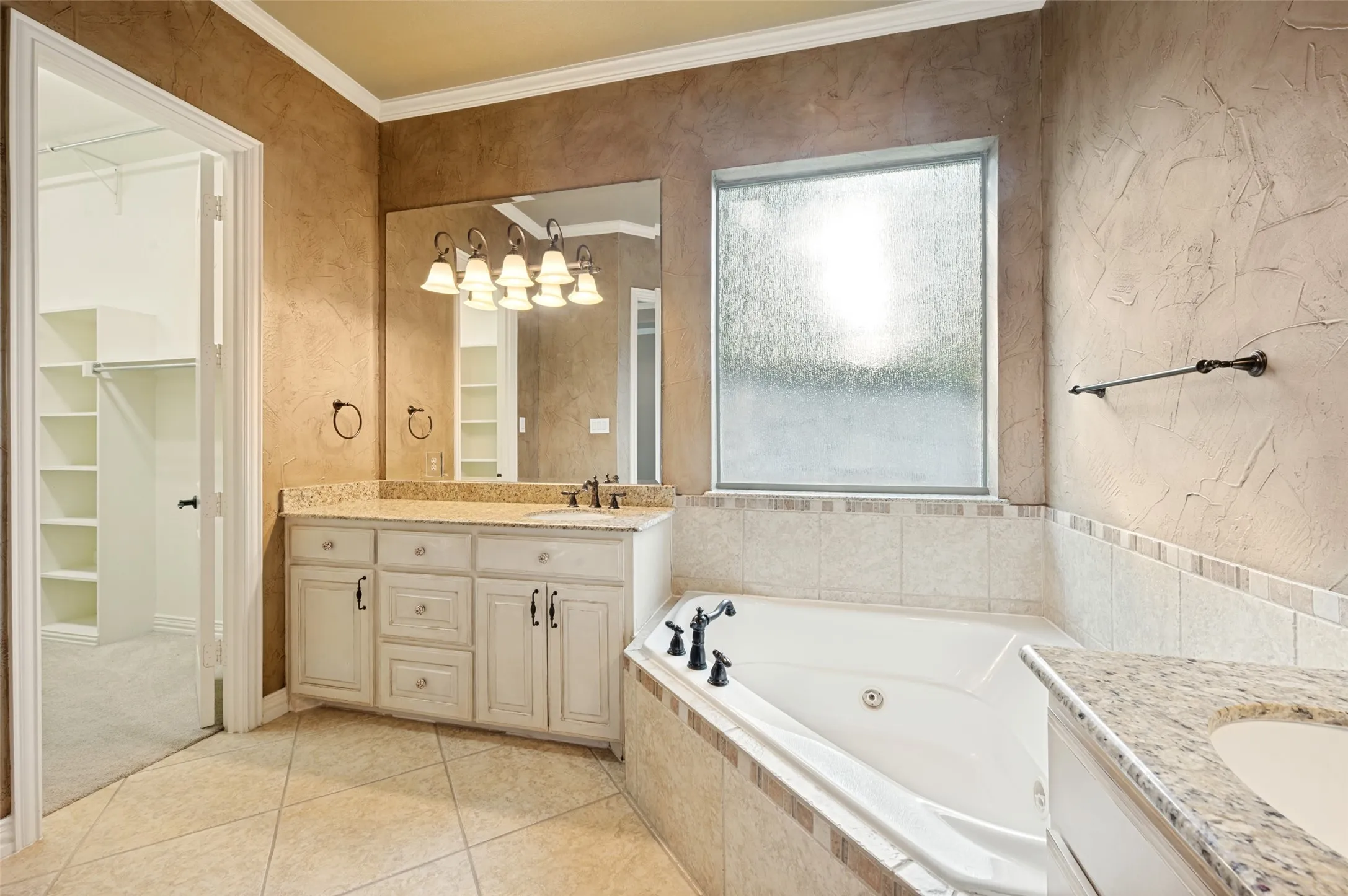 Soaking tub with tile surround, dual vanity with granite countertops, decorative wall texture, and tiled flooring