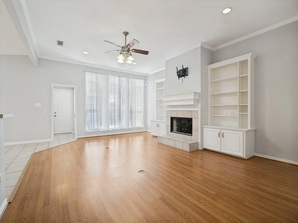 Spacious living area featuring wood-finish flooring, a tiled fireplace with mantel, built-in shelving, and large windows