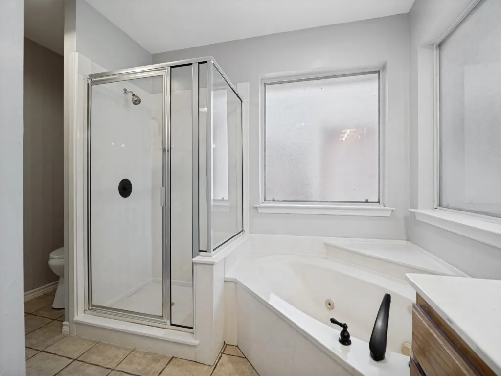 Tiled flooring, glass-enclosed shower, corner jetted tub with dark bronze fixtures, frosted windows, and a wood-finish vanity with a light-toned countertop