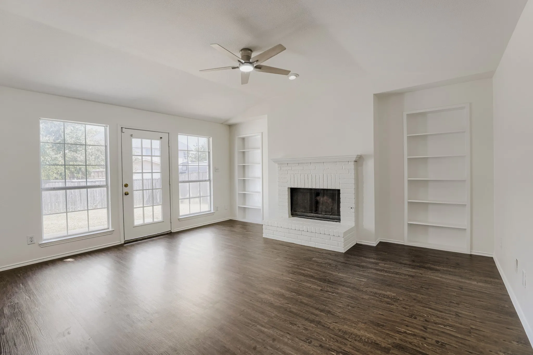 Vaulted ceiling with a ceiling fan, wood-finish flooring, two large windows, glass-paned door, and a brick fireplace
