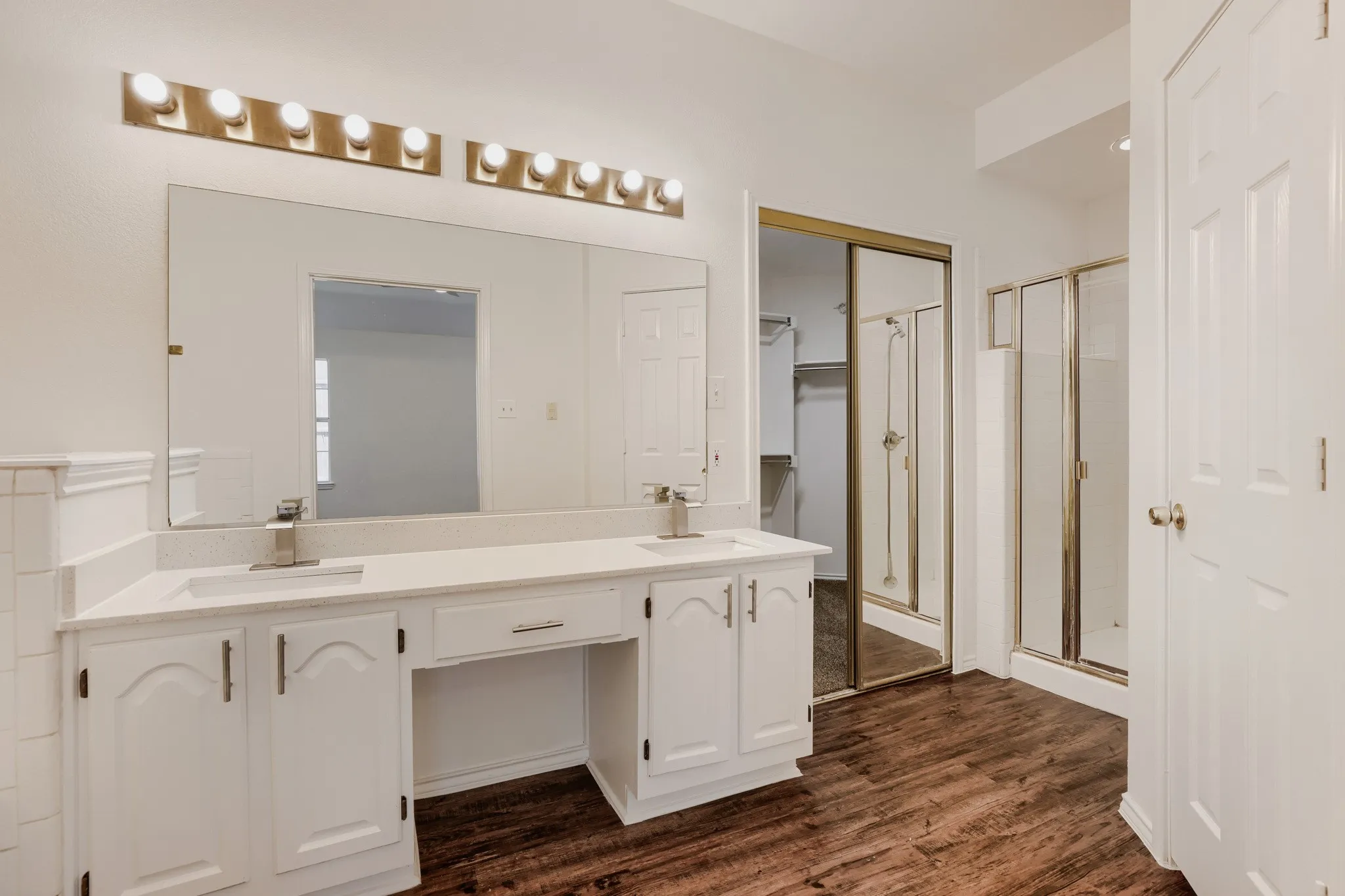 Dual vanity with individual sinks, white cabinetry, wood-finish flooring, and a glass-enclosed shower