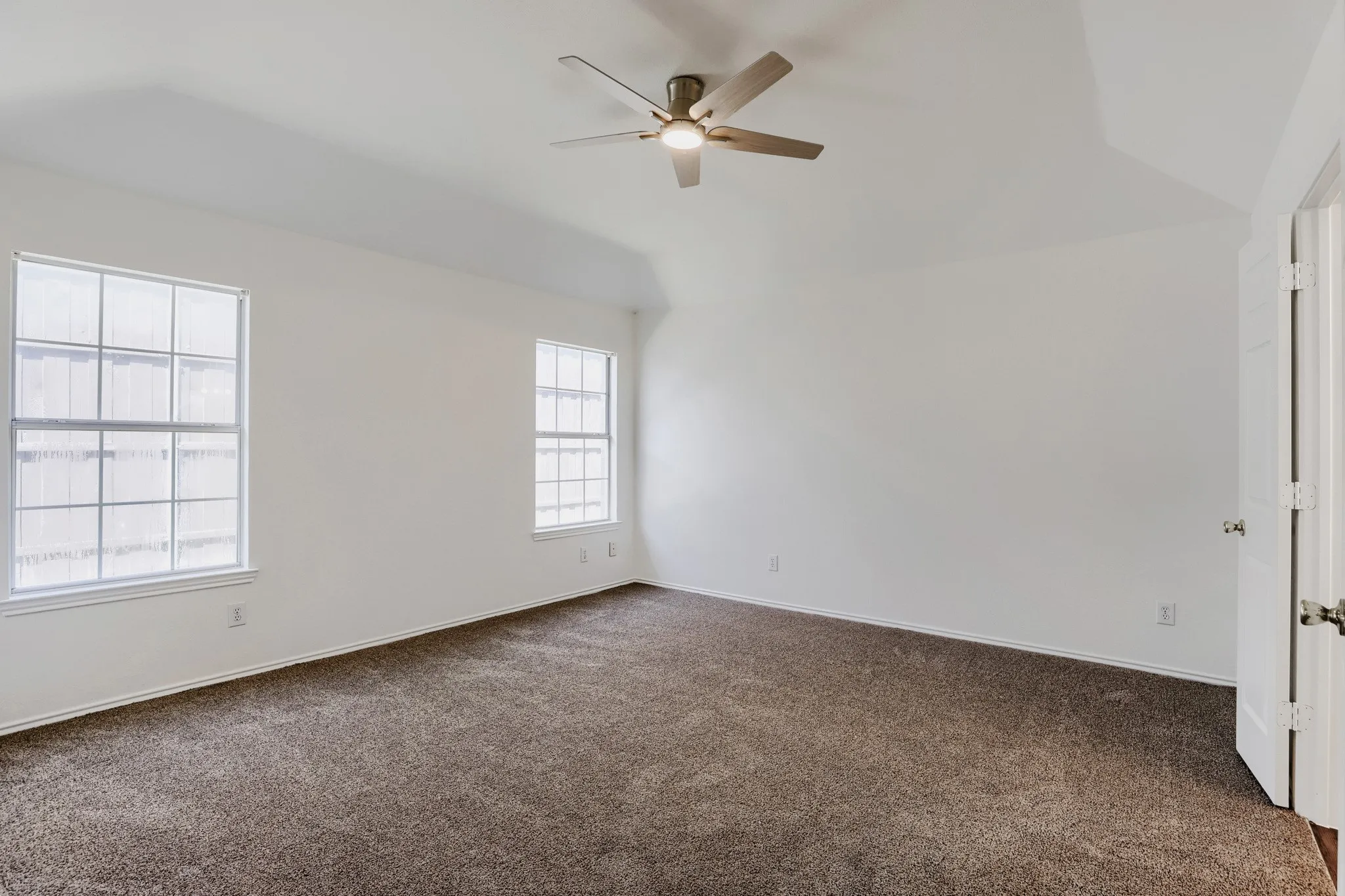 Spacious room featuring vaulted ceilings, a ceiling fan with integrated lighting, two multi-pane windows, neutral carpeting, and a white paneled door