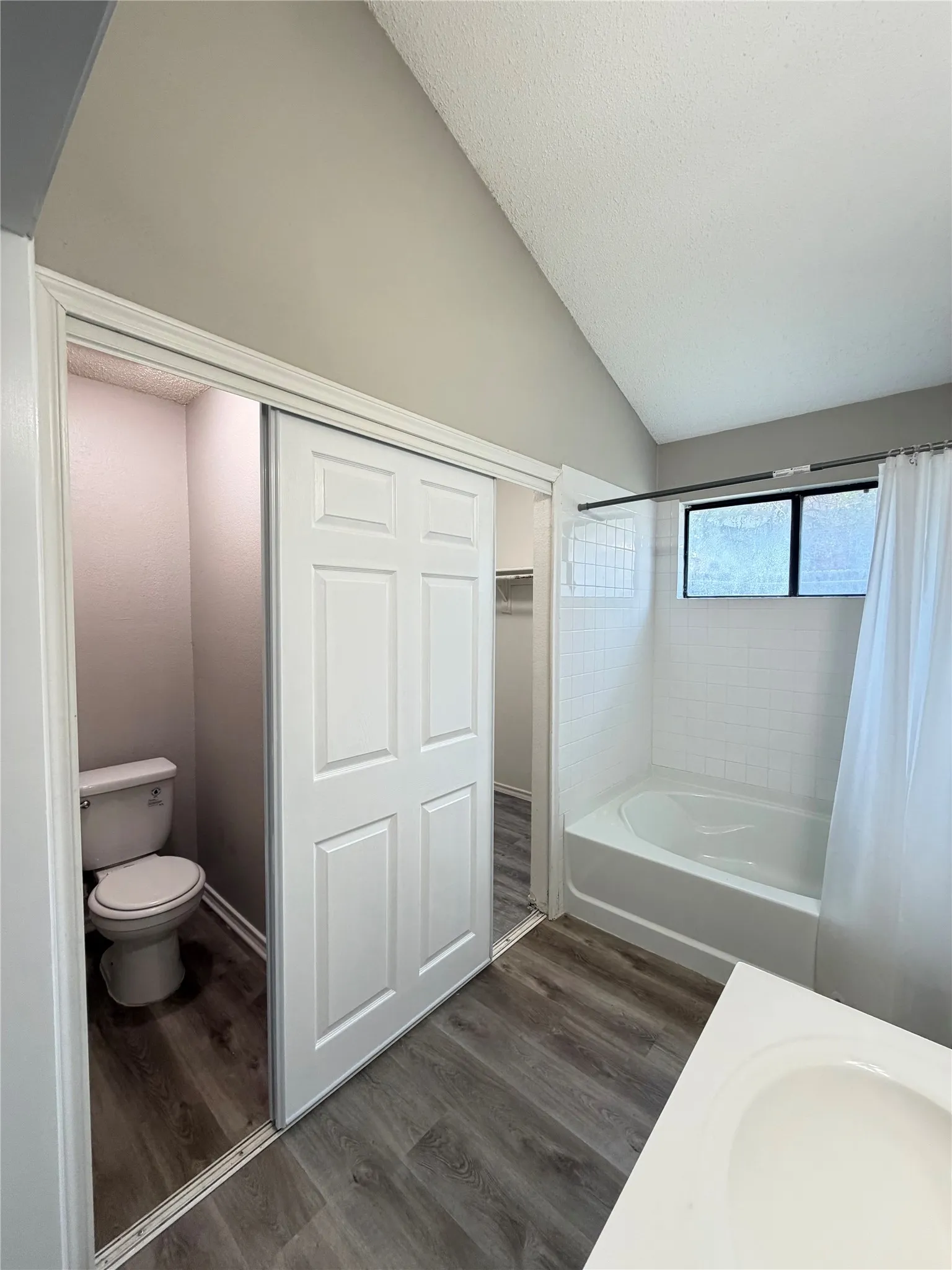 Vaulted ceiling, wood-finish flooring, white paneled bifold doors, white bathtub with tiled surround, and a vanity with an integrated sink