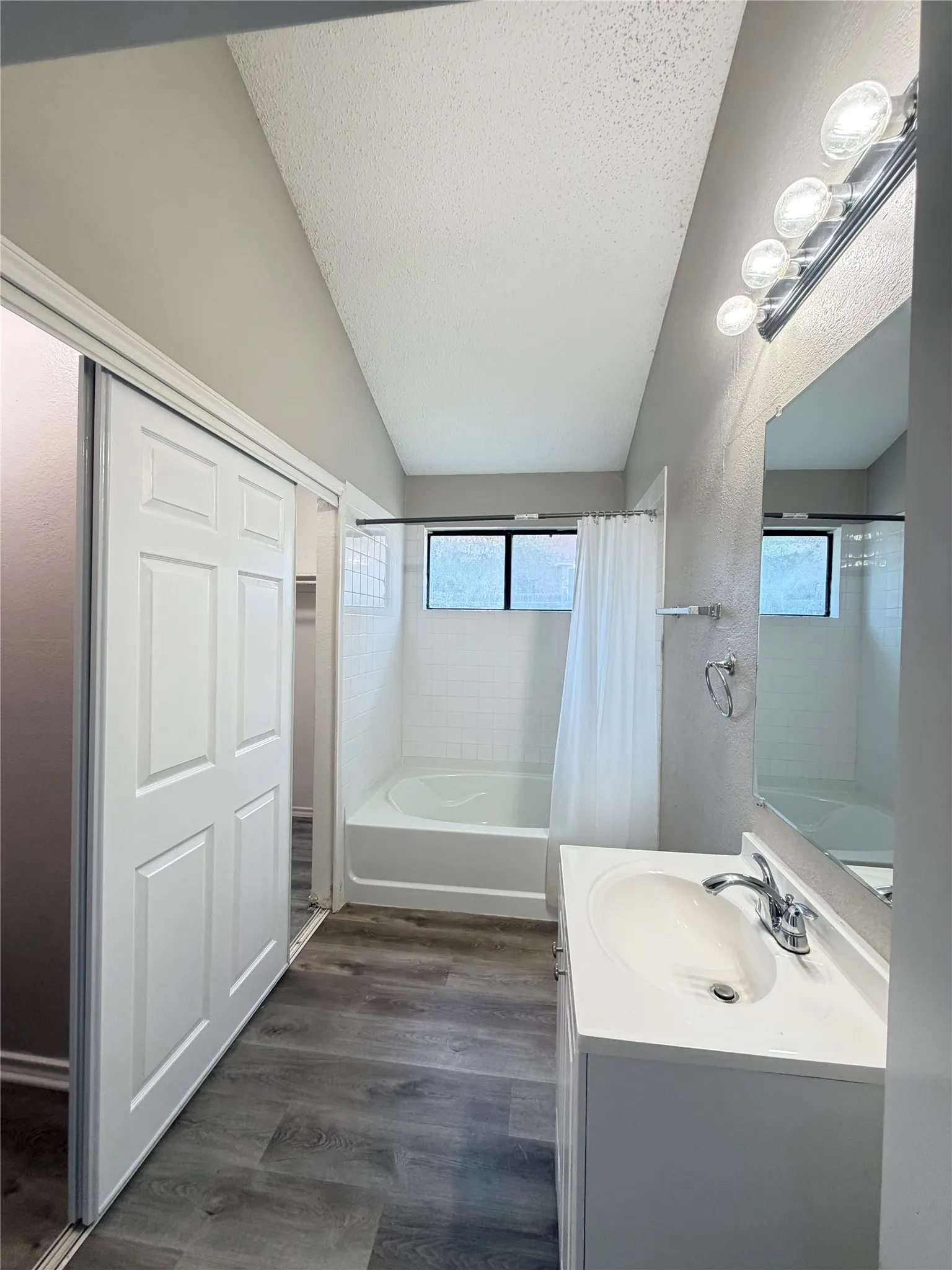 Vaulted ceiling, white panel closet doors, wood-finish flooring, white vanity with integrated sink, and a combination tub/shower with white subway tile surround