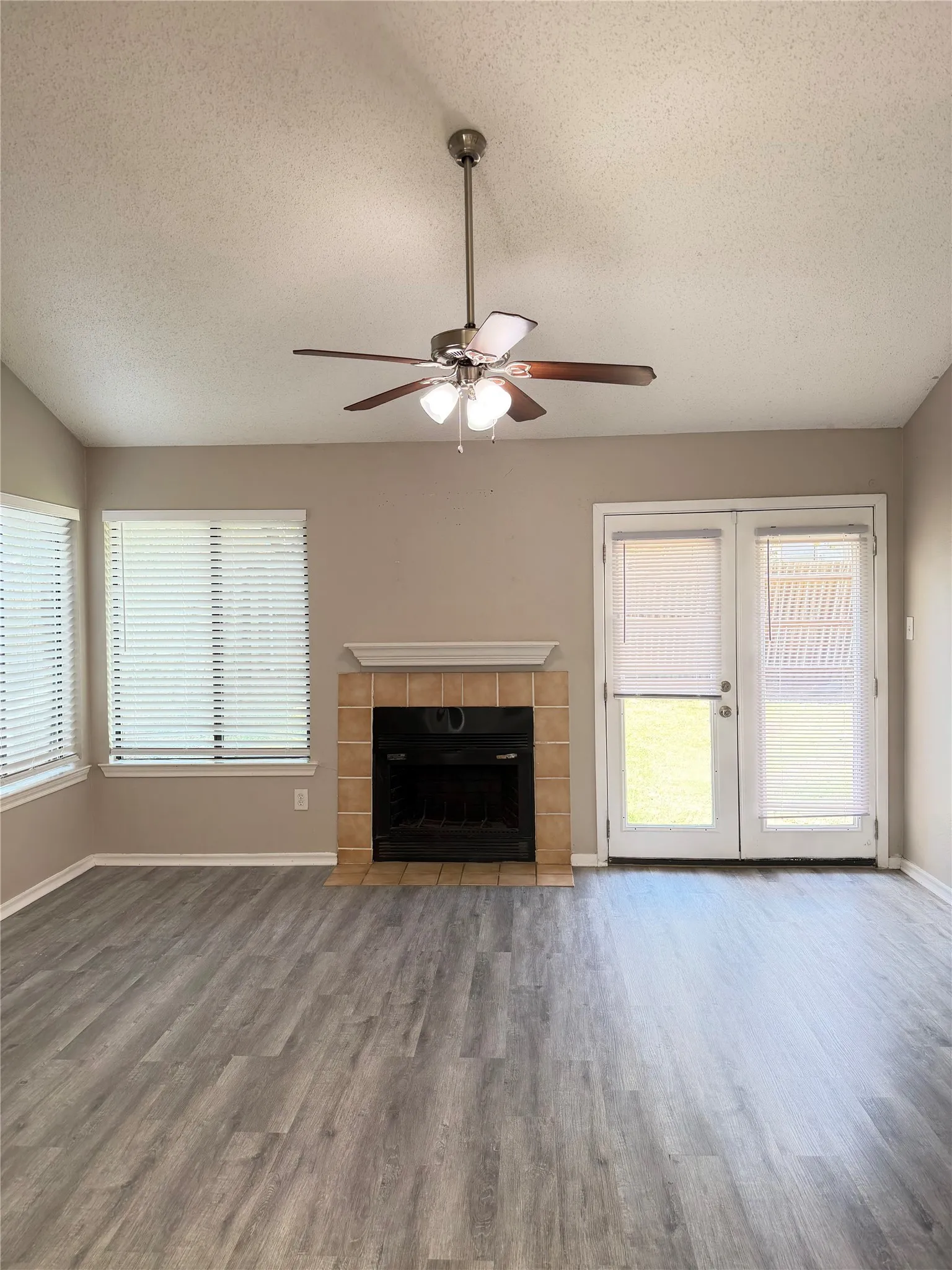 Wood-finish flooring, tiled fireplace with mantel, ceiling fan with integrated lighting, multiple windows with blinds, and white French doors