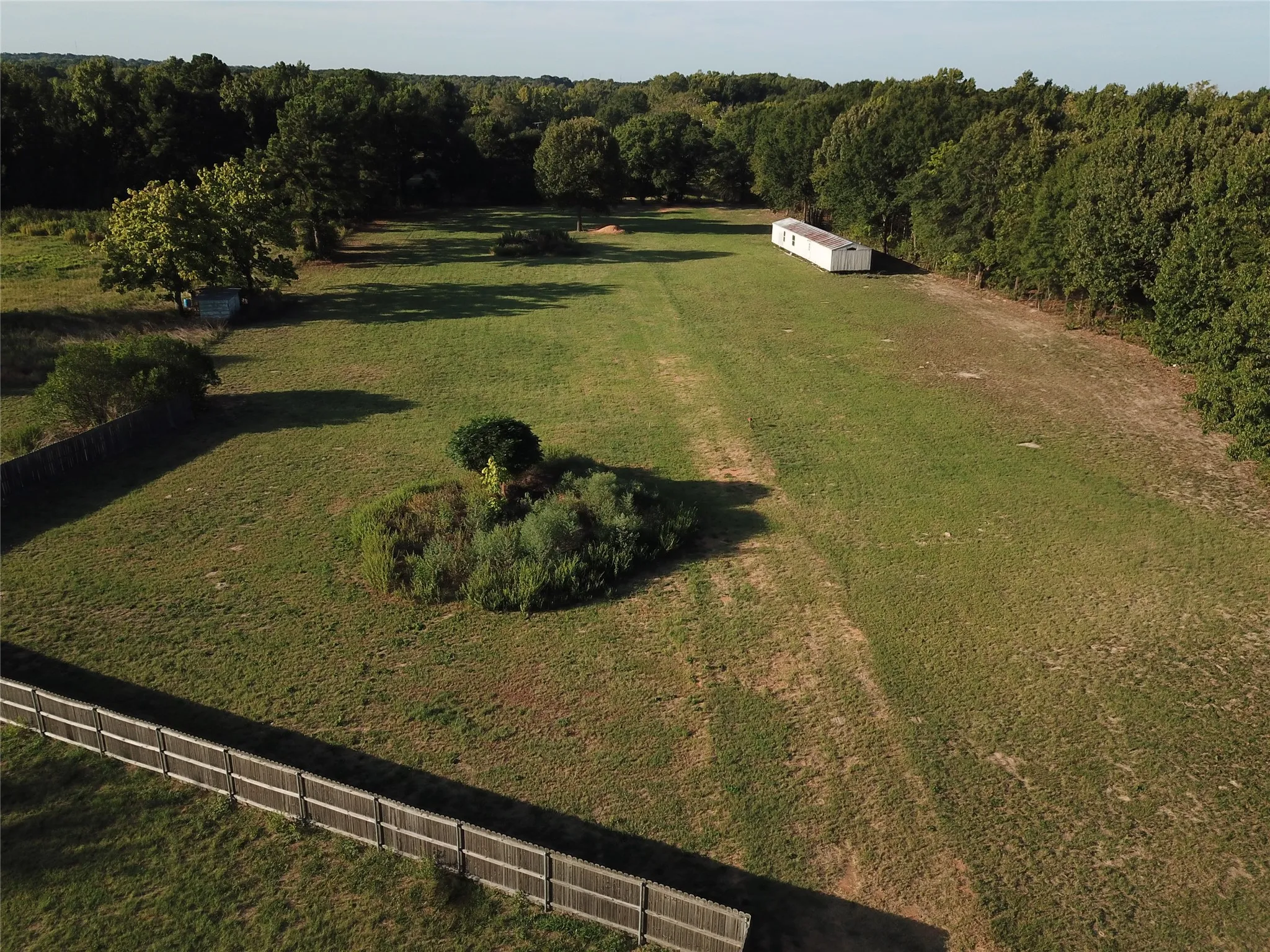 Expansive grassy lot featuring a perimeter fence, scattered mature trees, and a distant white structure