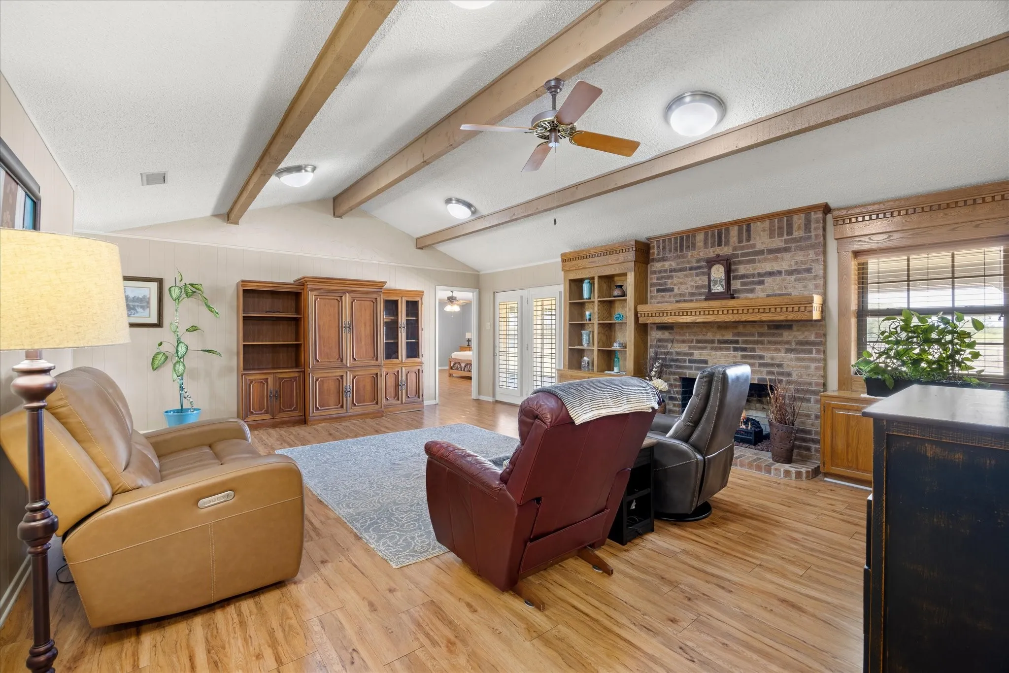 Vaulted ceiling with exposed wood beams, brick fireplace with wood mantel, wood-finish flooring, built-in shelving, and plantation shutters