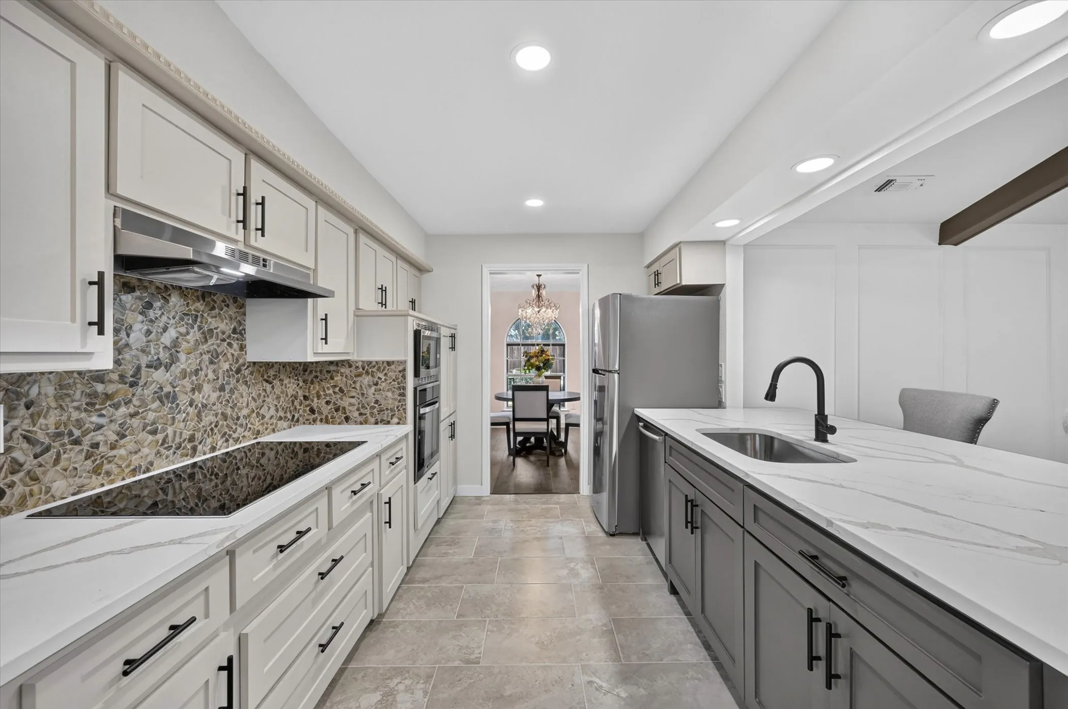 Galley kitchen featuring white upper cabinetry, dark lower cabinetry, stone backsplash, integrated cooktop, and stainless steel appliances