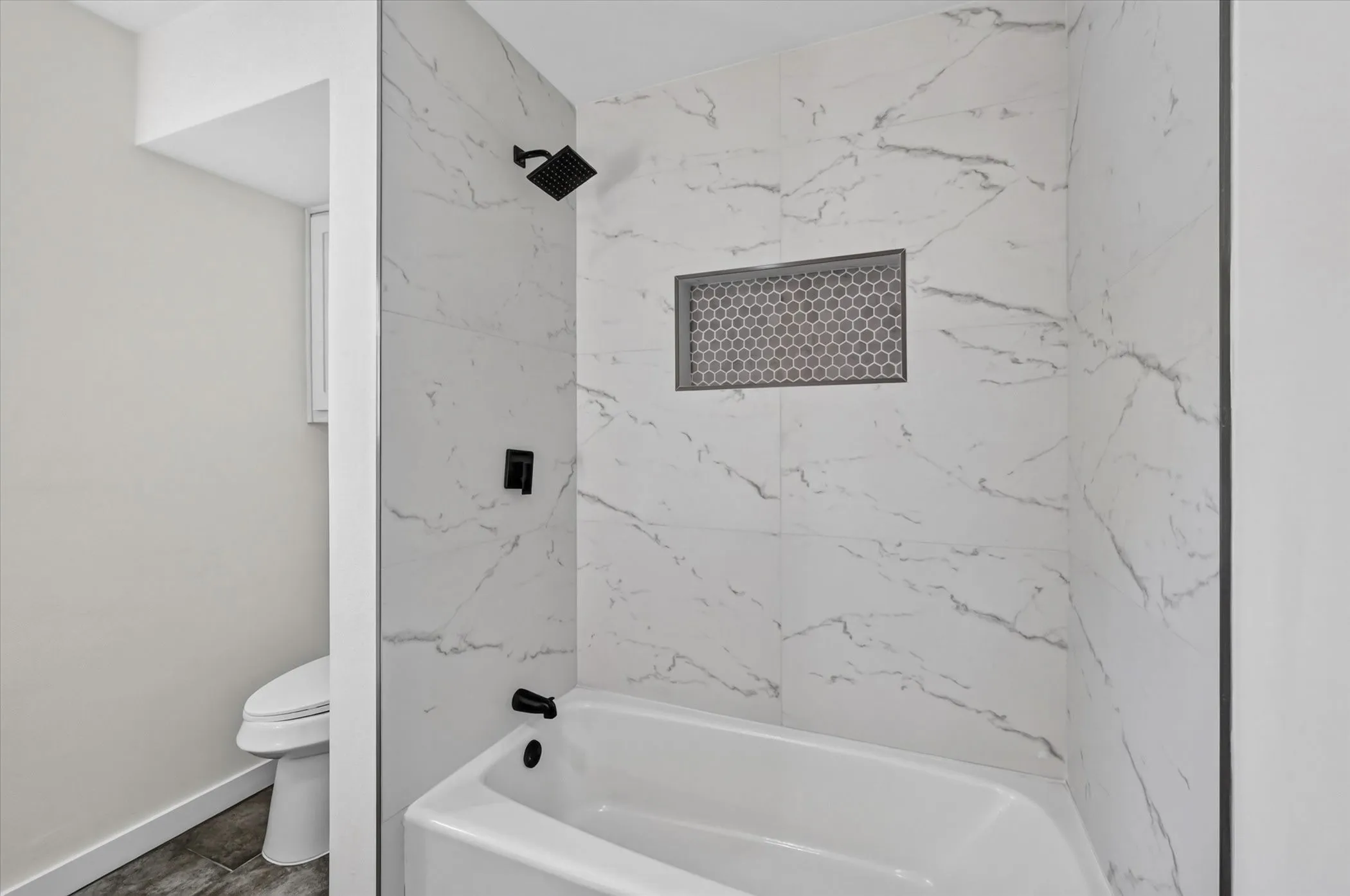 White tiled shower surround with dark grout, matte black shower fixtures, recessed shower niche with honeycomb mosaic tile, and a white soaking tub
