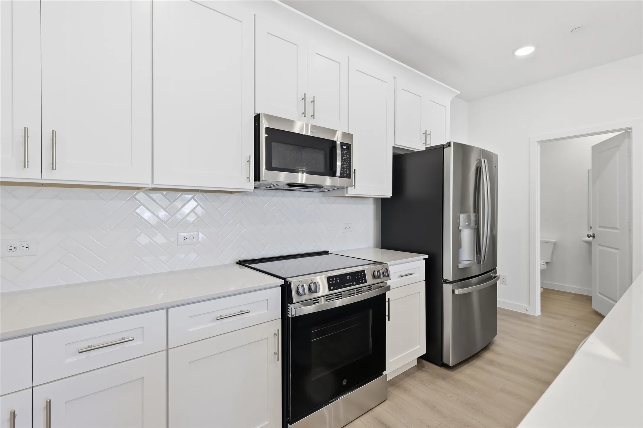 White shaker cabinetry with brushed nickel hardware, stainless steel appliances, white herringbone tile backsplash, and light wood-finish flooring