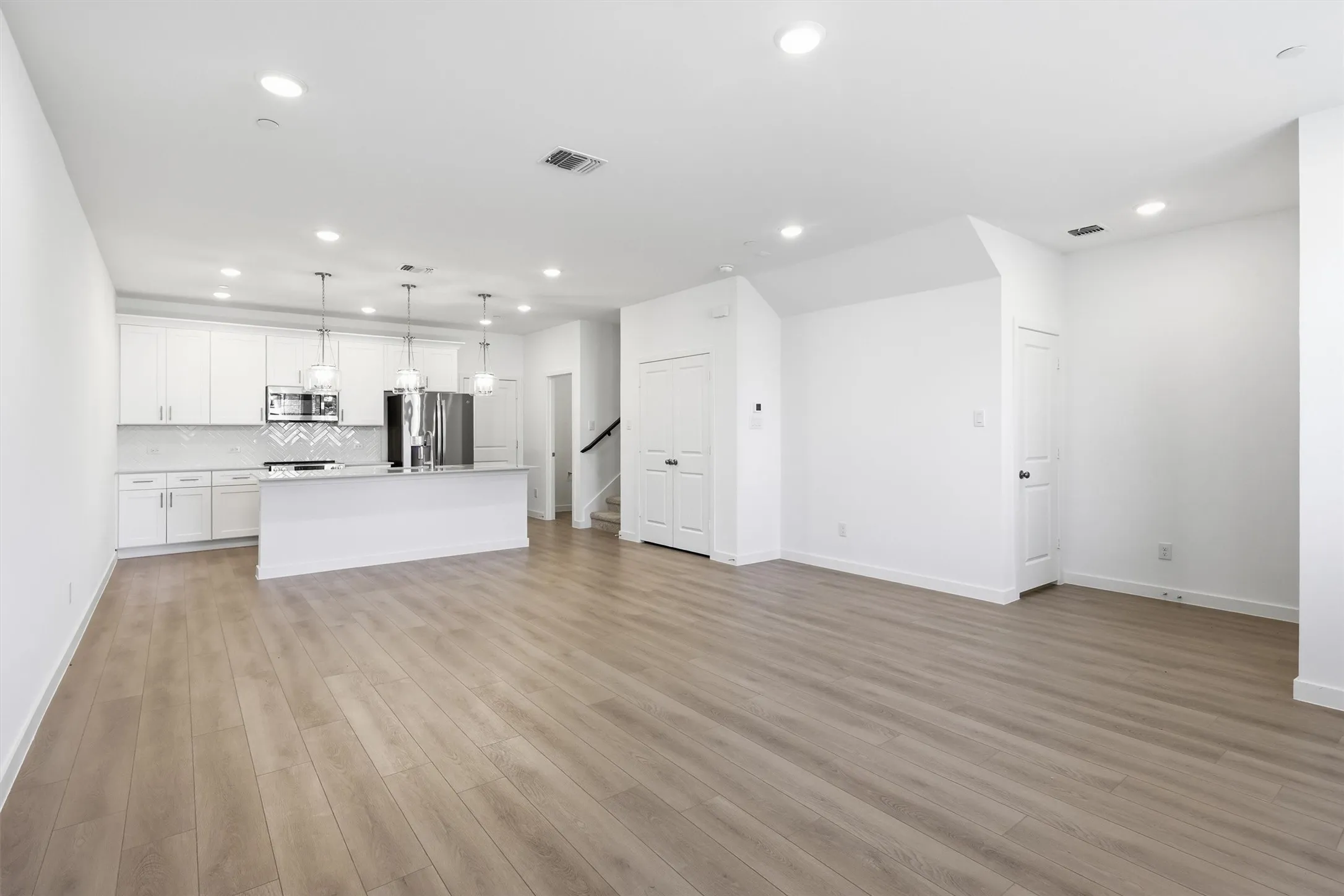 Open-concept living area with wood-finish flooring, recessed lighting, and a white kitchen featuring stainless steel appliances, a white tile backsplash, and a central island with pendant lighting