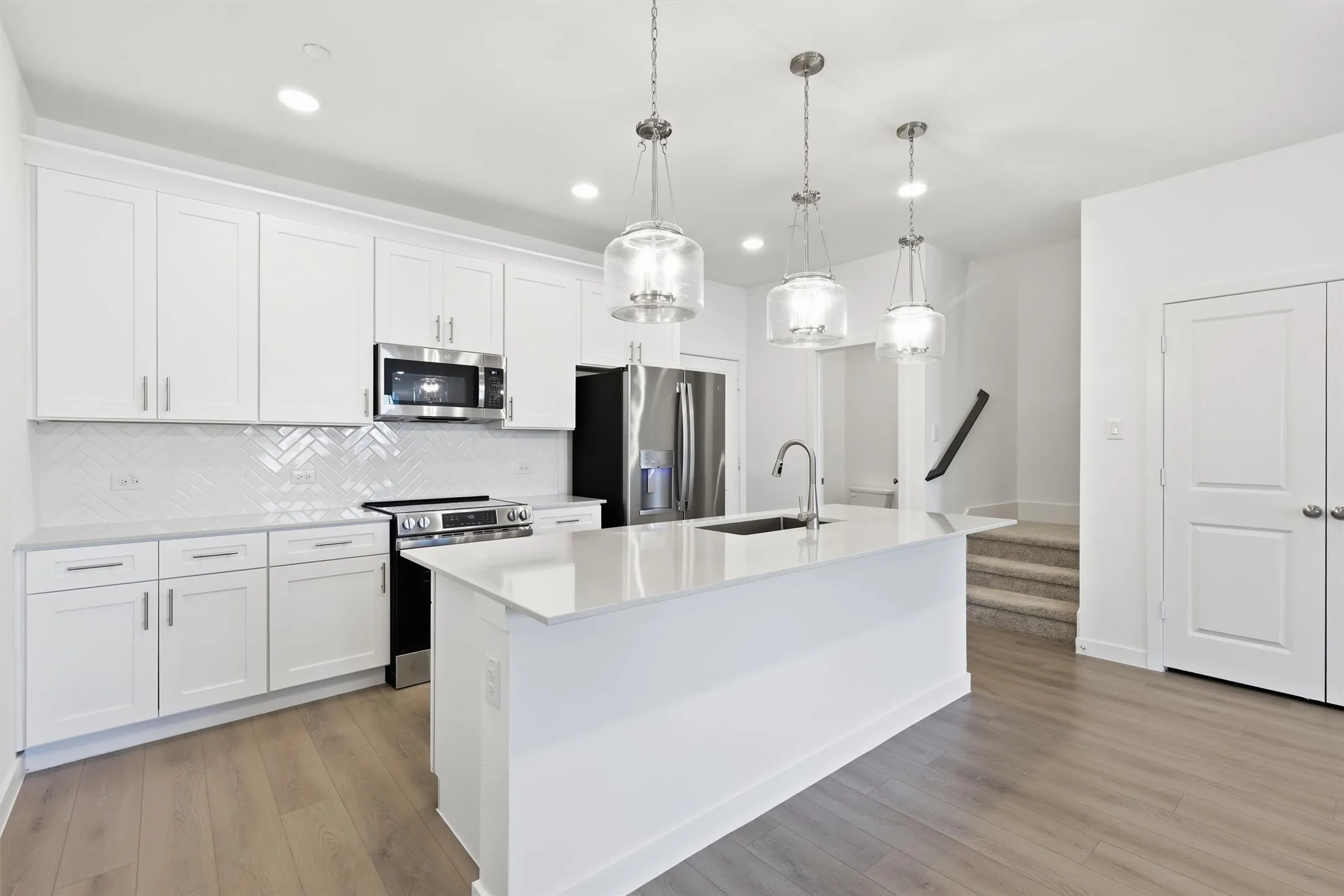 Kitchen featuring white shaker cabinetry, stainless steel appliances, a herringbone tile backsplash, an island with an undermount sink, and wood-finish flooring