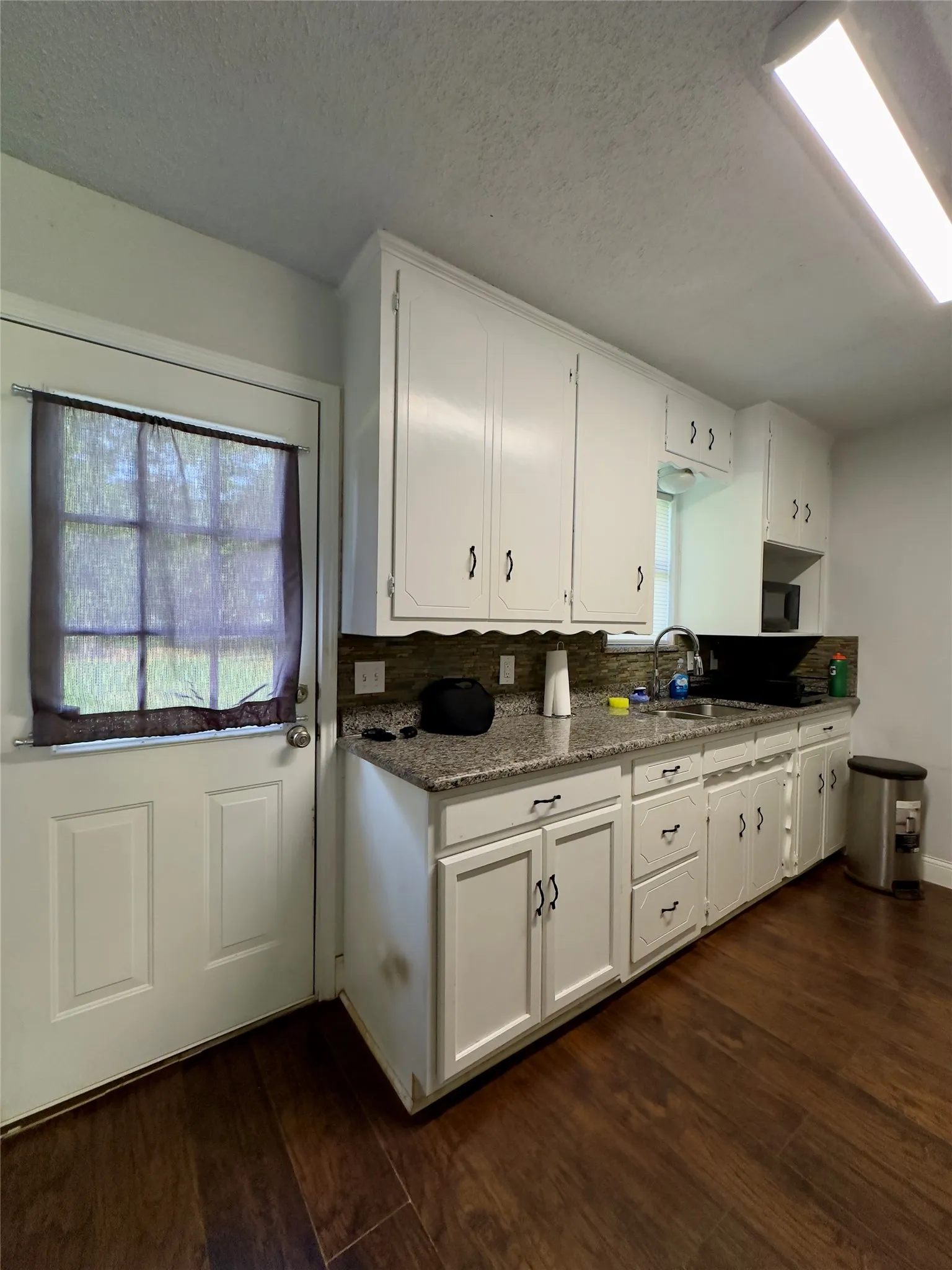White cabinetry, granite-finish countertops, tile backsplash, stainless steel sink, and wood-finish flooring