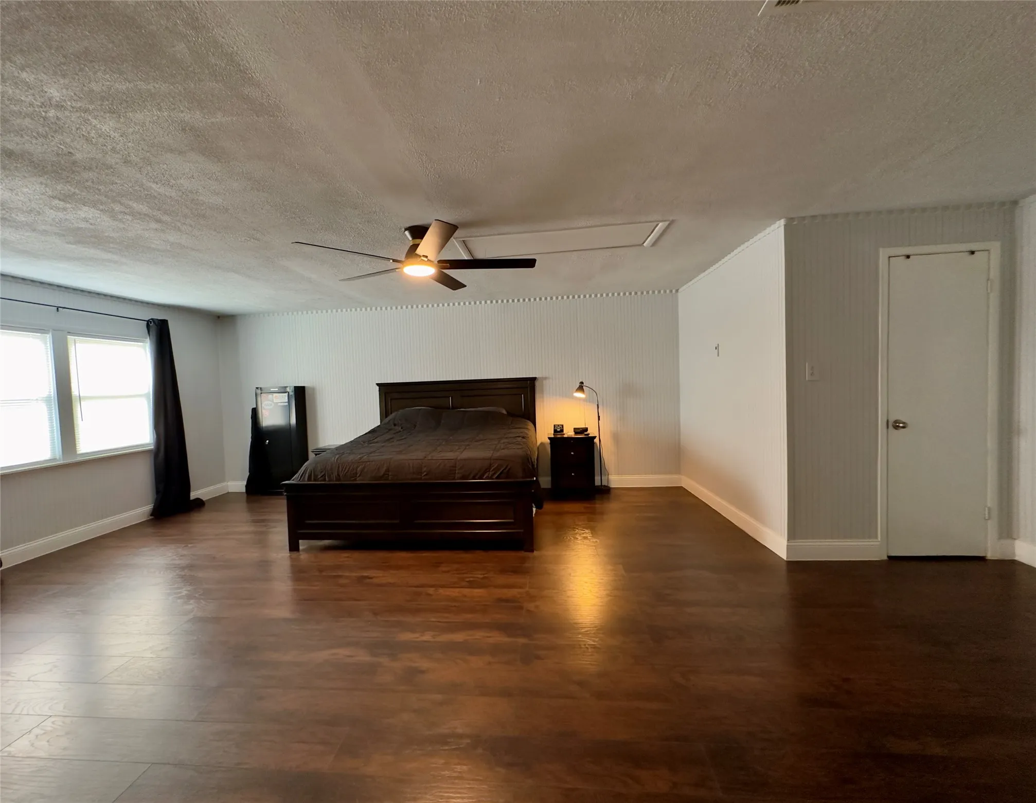 Spacious room featuring wood-finish flooring, multiple windows, a ceiling fan with integrated lighting, and a white interior door