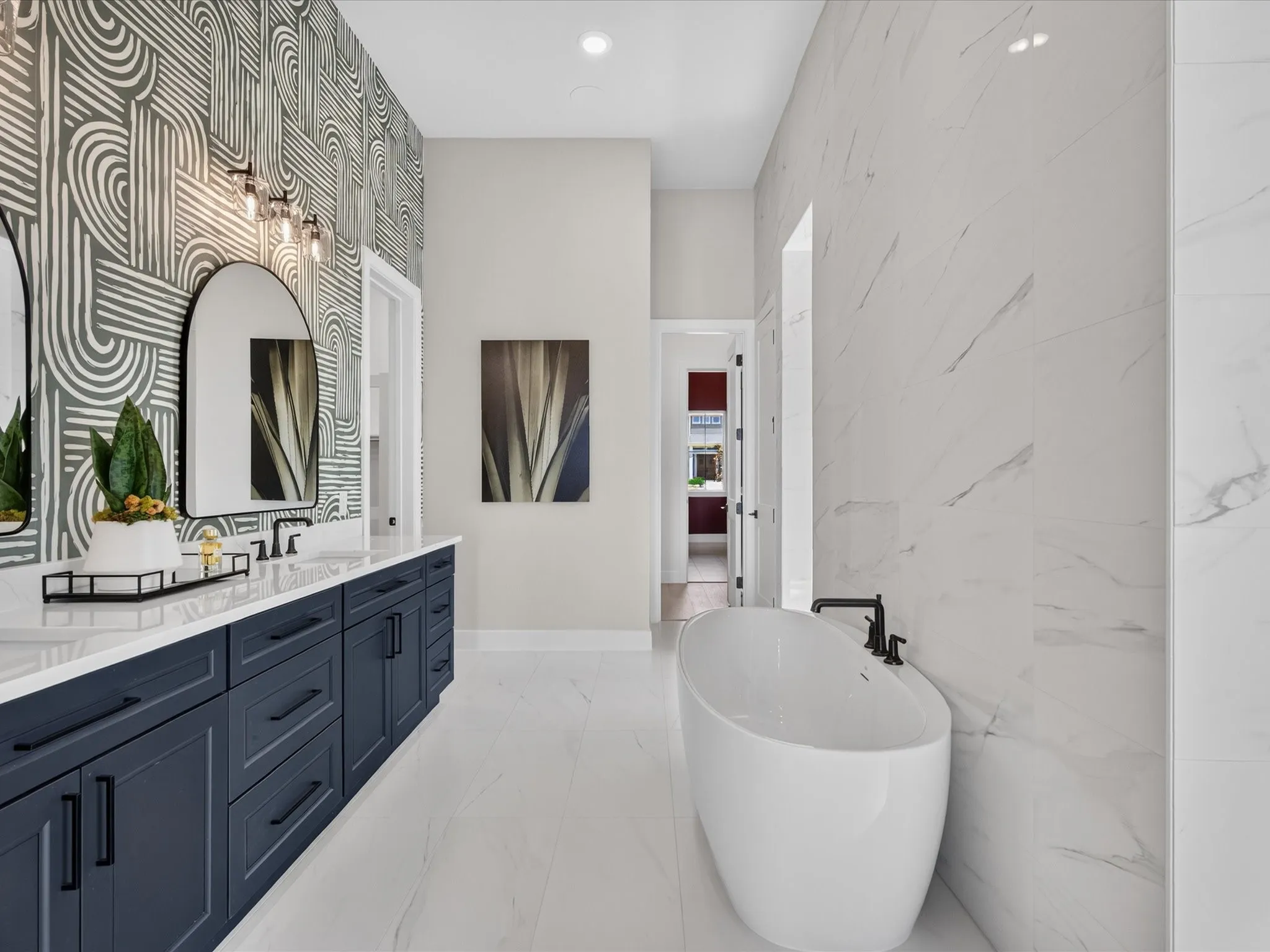 Freestanding soaking tub, dual vanity with undermount sinks, patterned accent wall, white marble-look floor tile, and white marble-look wall tile