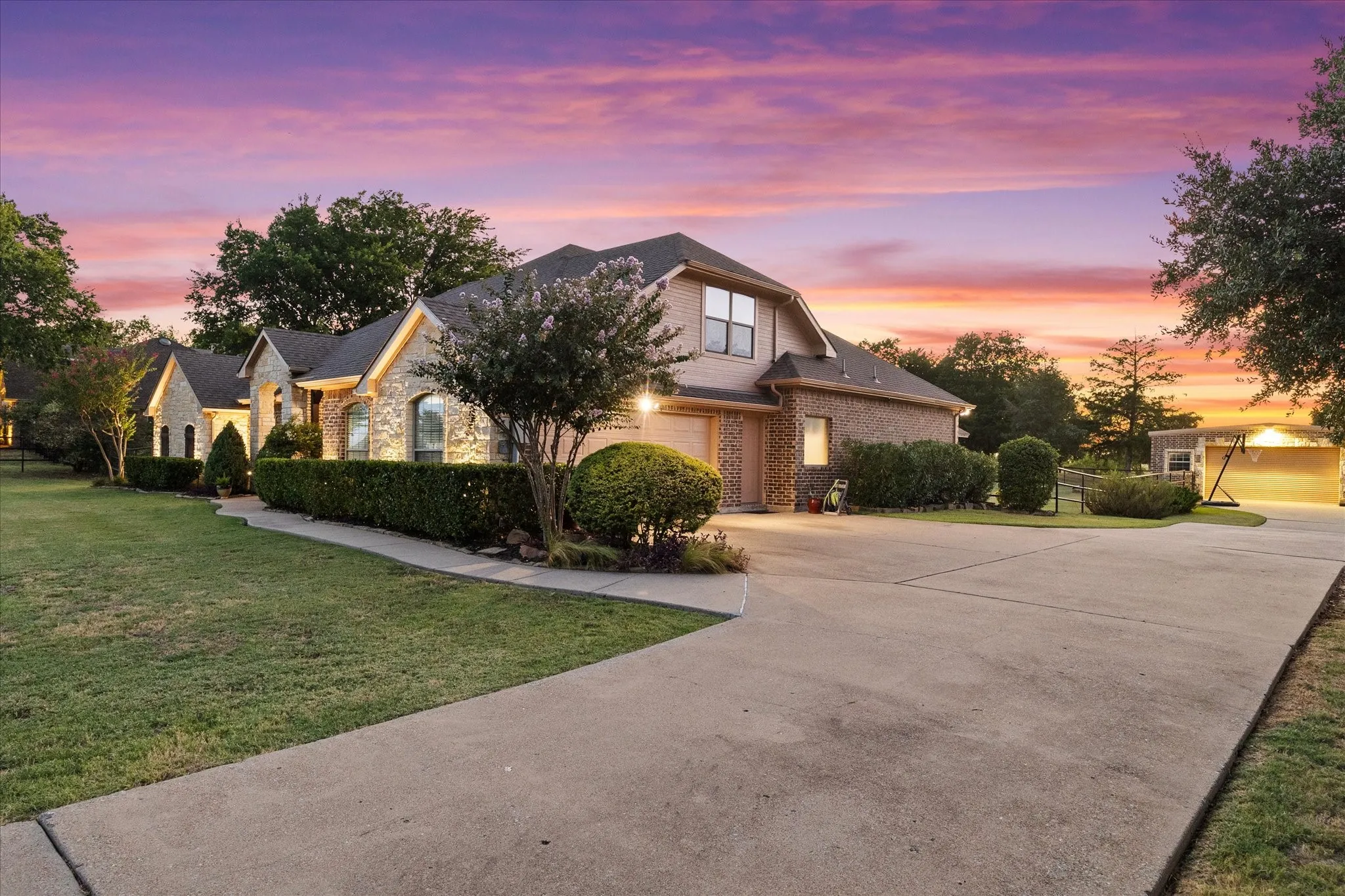 Stone and brick exterior with a multi-level design, concrete driveway, manicured landscaping, and mature trees