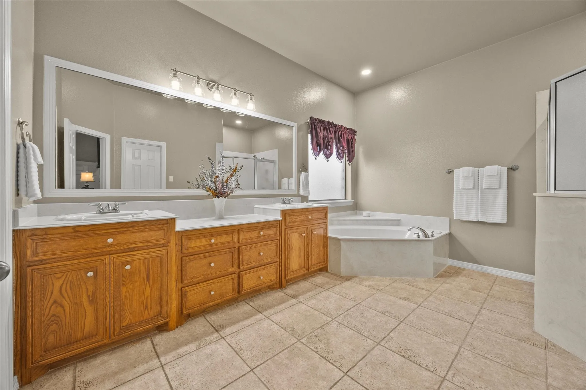 Dual wood-finish vanities with white countertops, expansive wall-mounted mirror, inset corner soaking tub, and tiled flooring