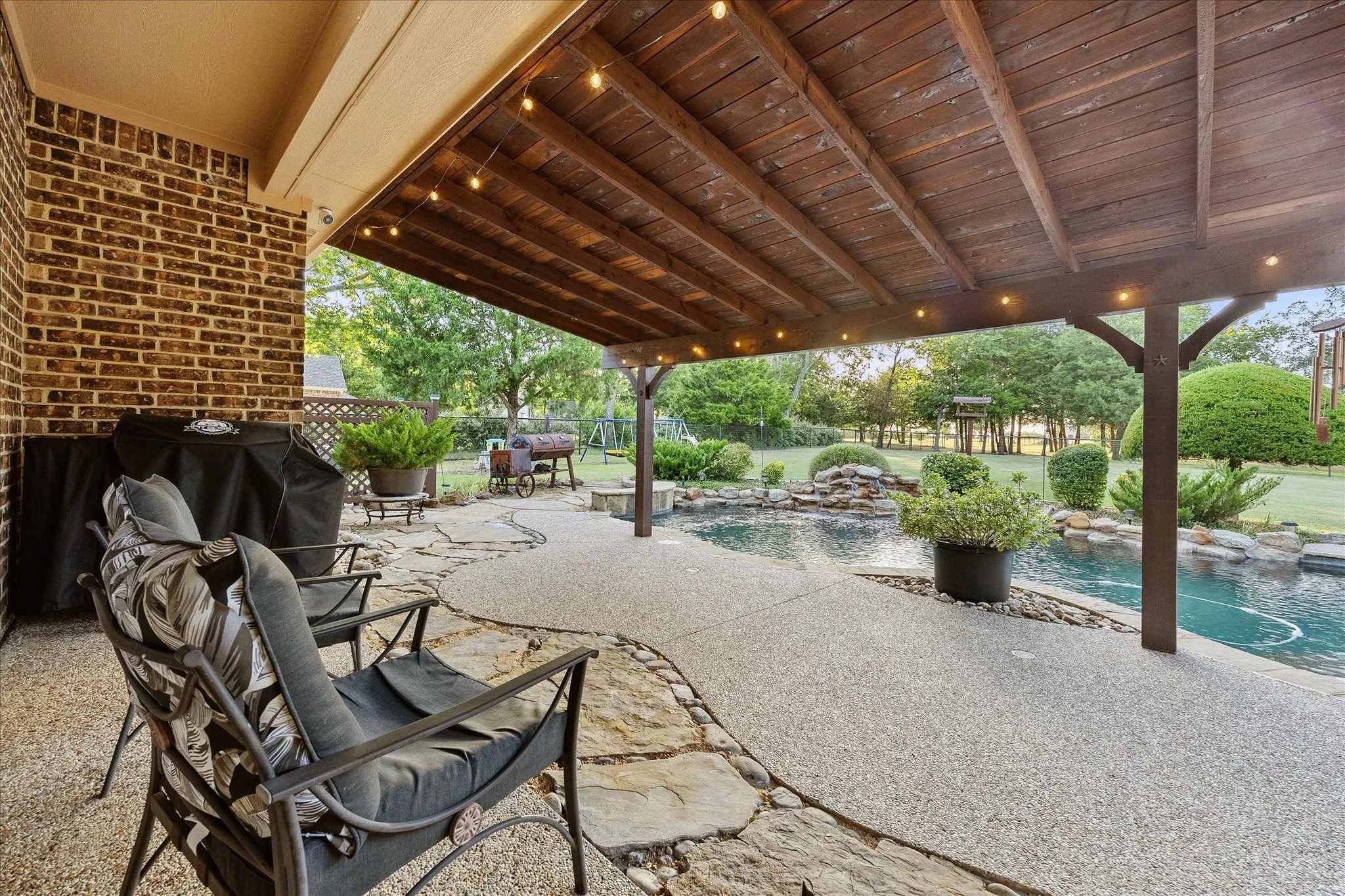 Covered patio with exposed aggregate flooring and brick wall, featuring an adjacent swimming pool with a rock waterfall