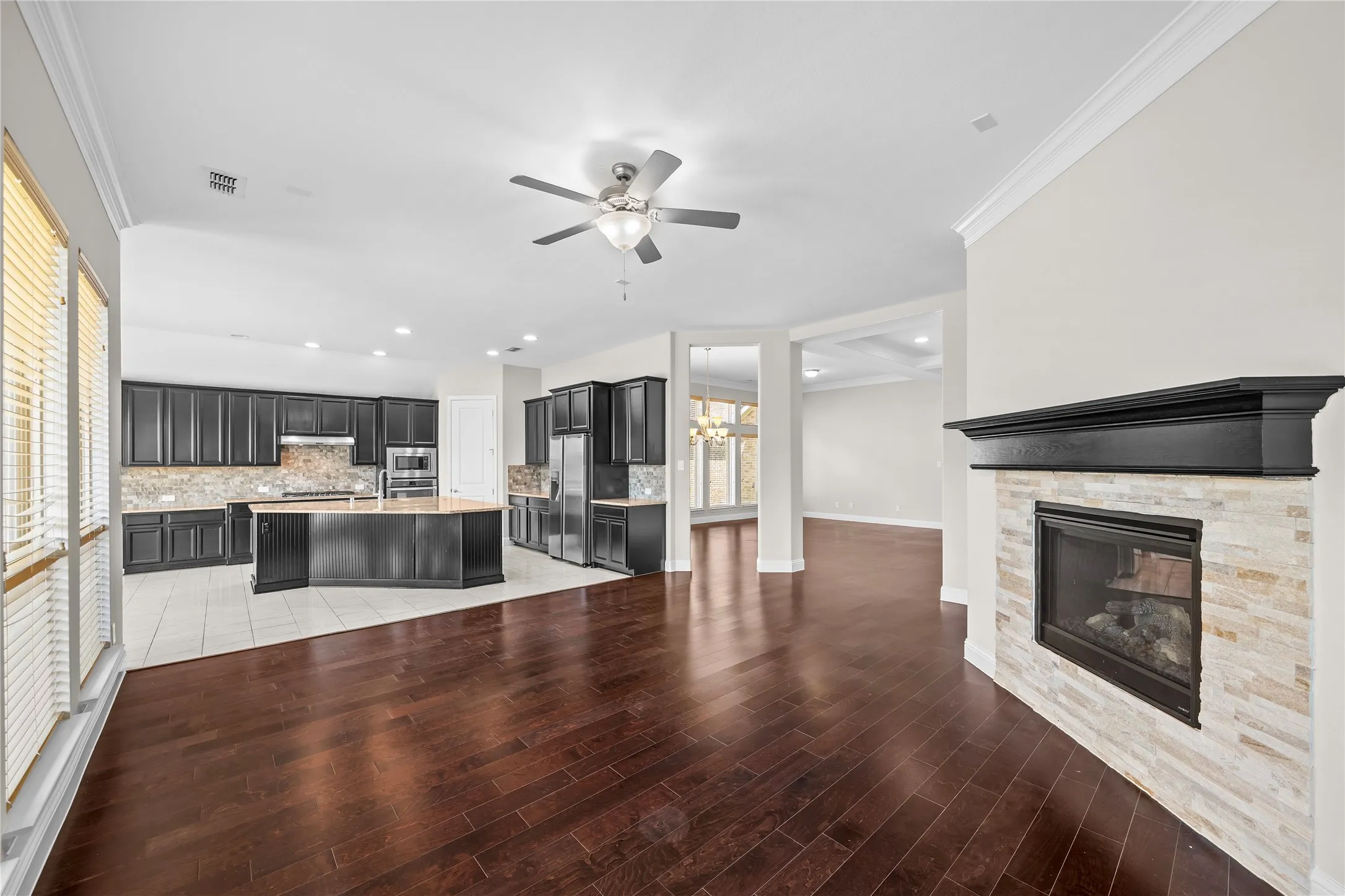 Open-concept living area with dark wood-finish flooring, a stone-clad fireplace with a dark mantel, and a kitchen featuring dark cabinetry, an island, and stainless steel appliances