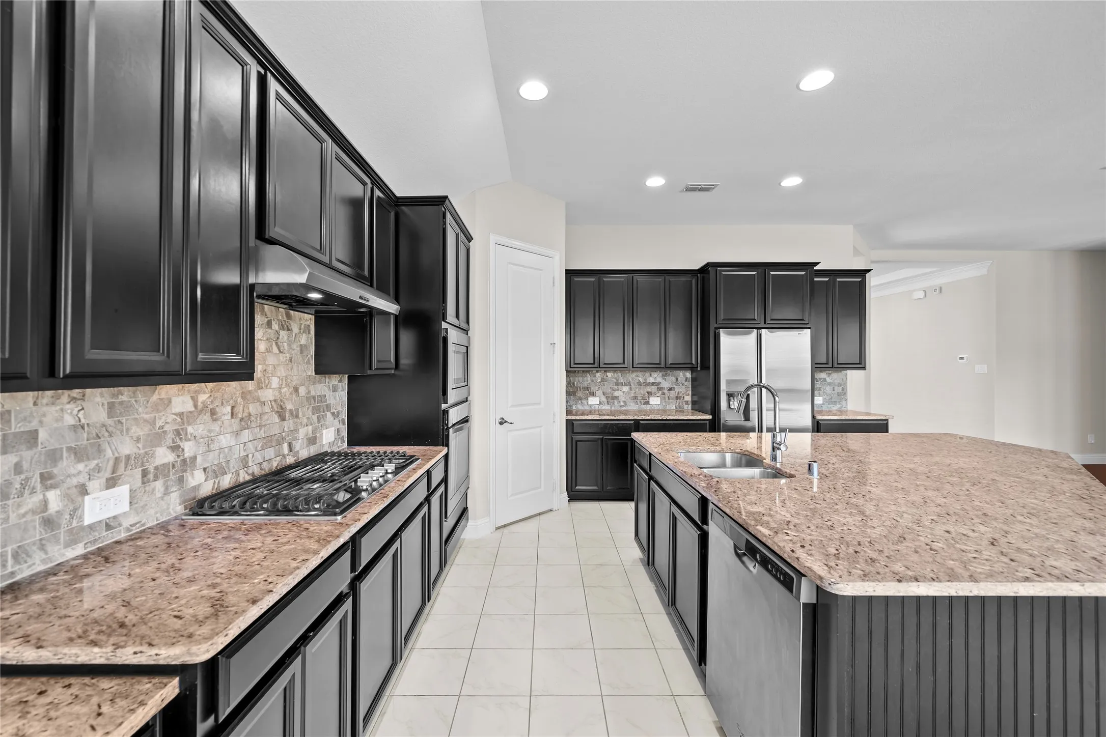 Spacious kitchen featuring dark wood cabinetry, light tile flooring, an oversized island with dual sinks, built-in stainless steel appliances, and a gas cooktop