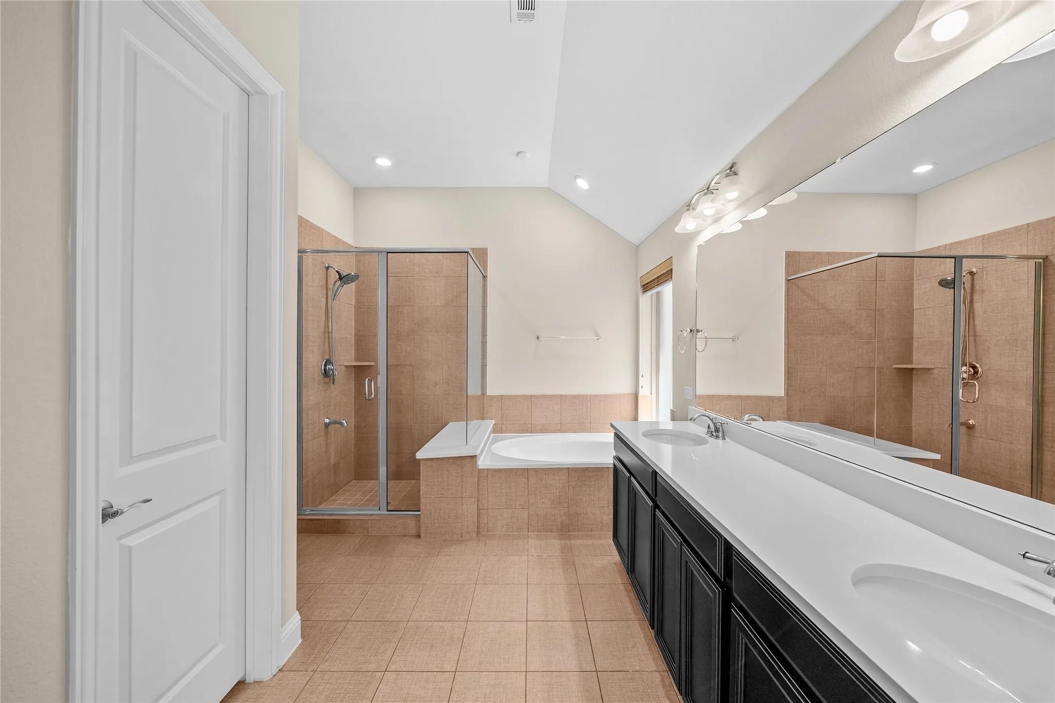 Spacious bathroom featuring a dual vanity with undermount sinks, tiled flooring, a glass-enclosed shower, a built-in bathtub, and a vaulted ceiling