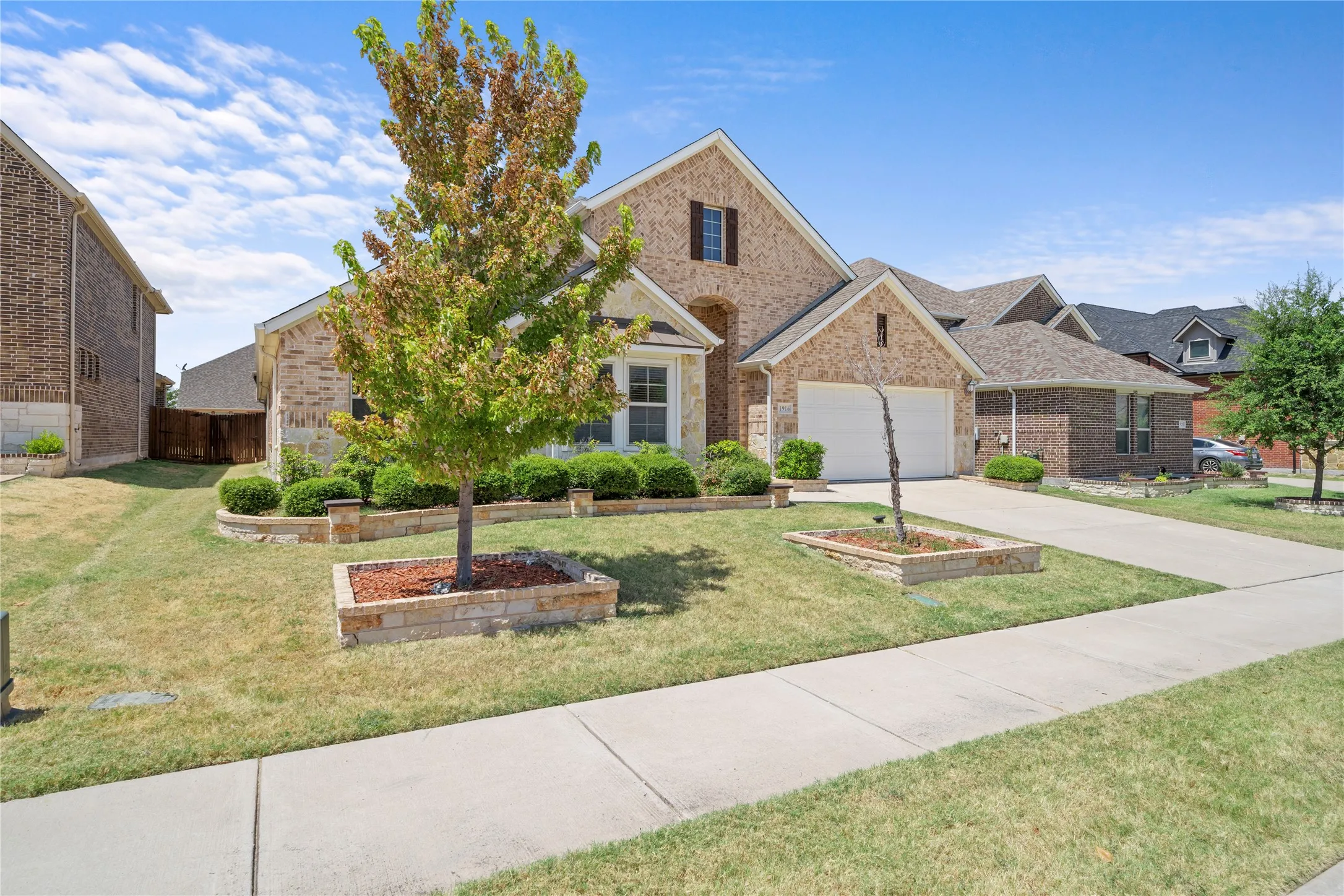Brick exterior with stone accents, two-car garage, concrete driveway, front yard landscaping, and a sidewalk