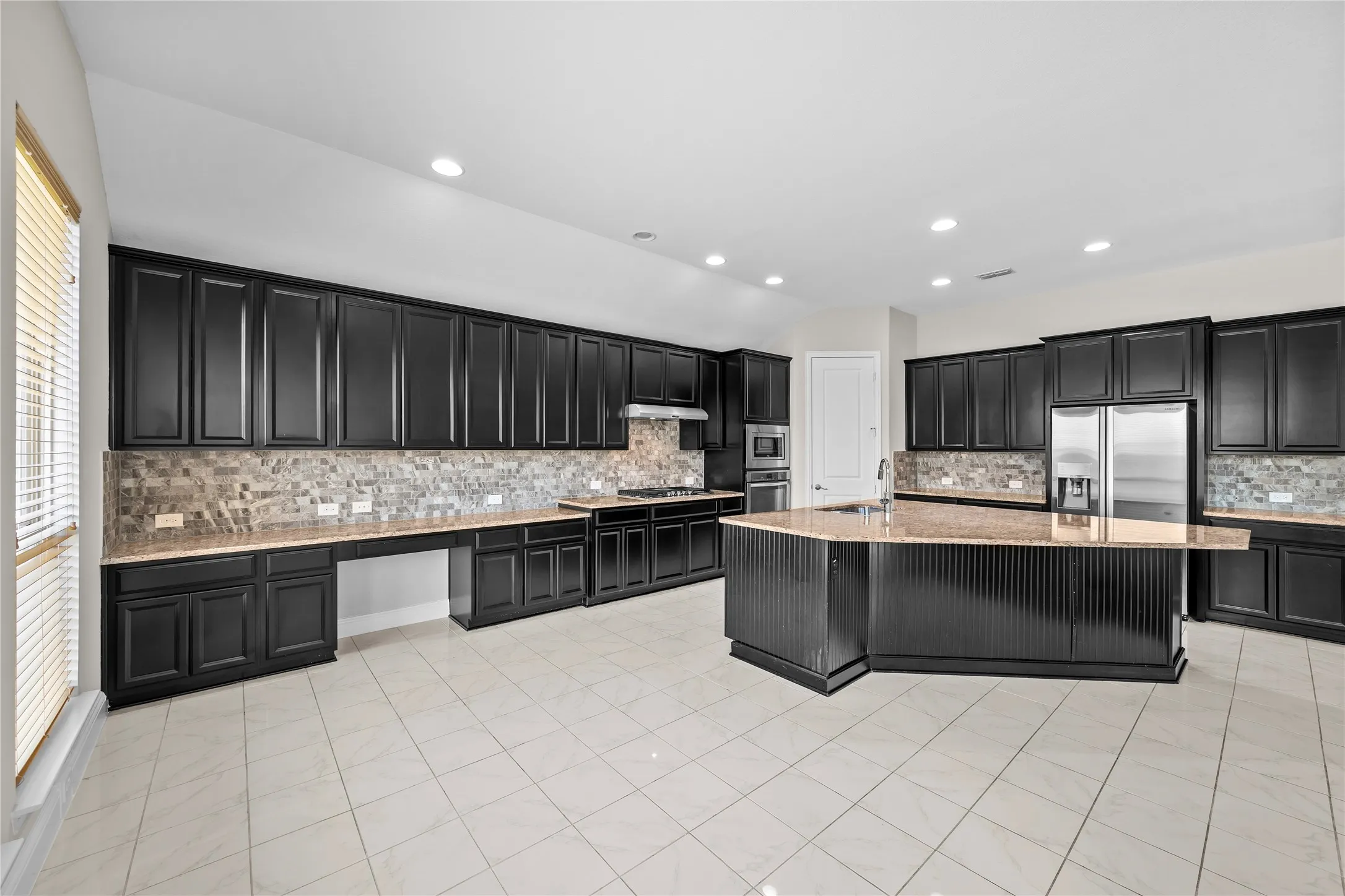 Spacious kitchen featuring extensive dark wood cabinetry, light-toned tile flooring, a large multi-sided island with an integrated sink, stone countertops, and a full stainless steel appliance suite
